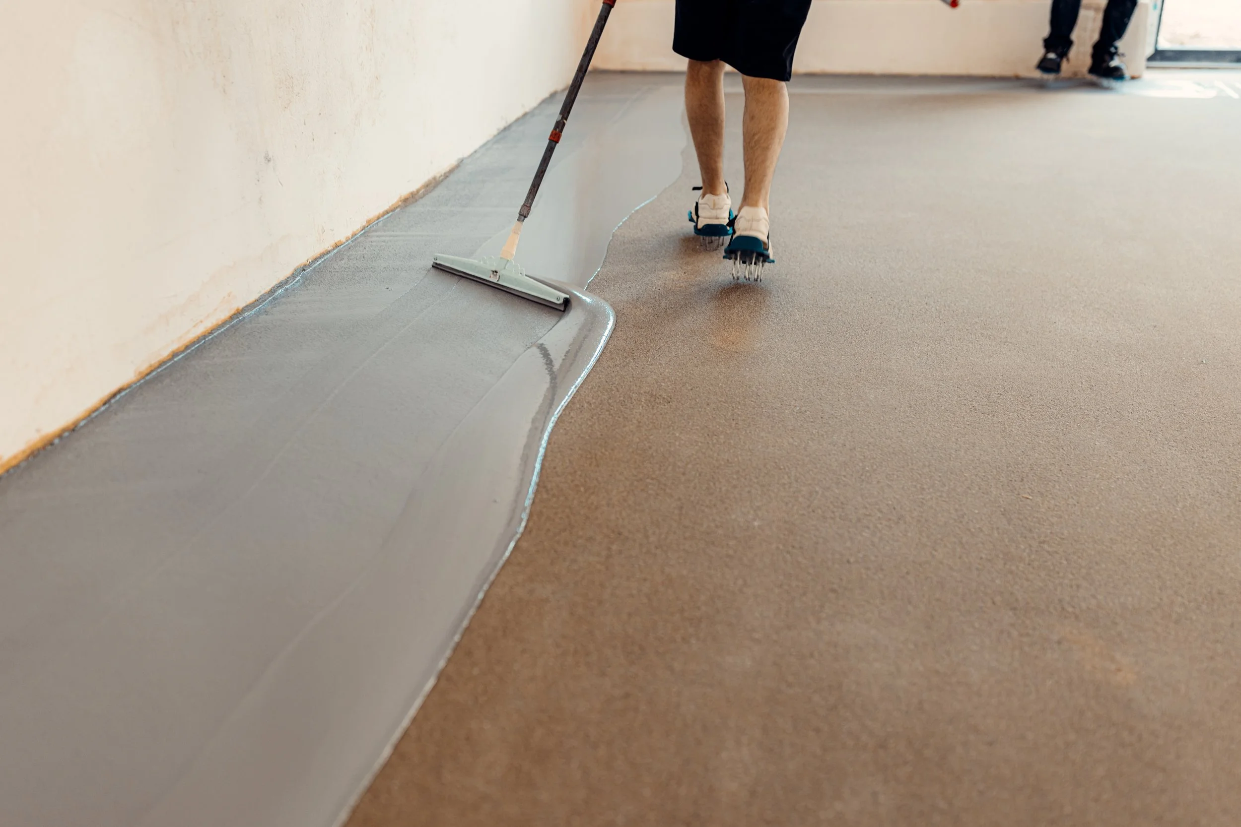 Person applying a gray coating to a brown floor using a squeegee, wearing roller skates.