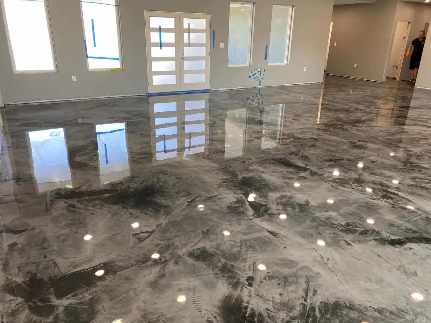Interior view of a room with polished gray marble flooring, unfinished walls with blue tape, and a door leading outside. There is a person standing in a doorway on the right.