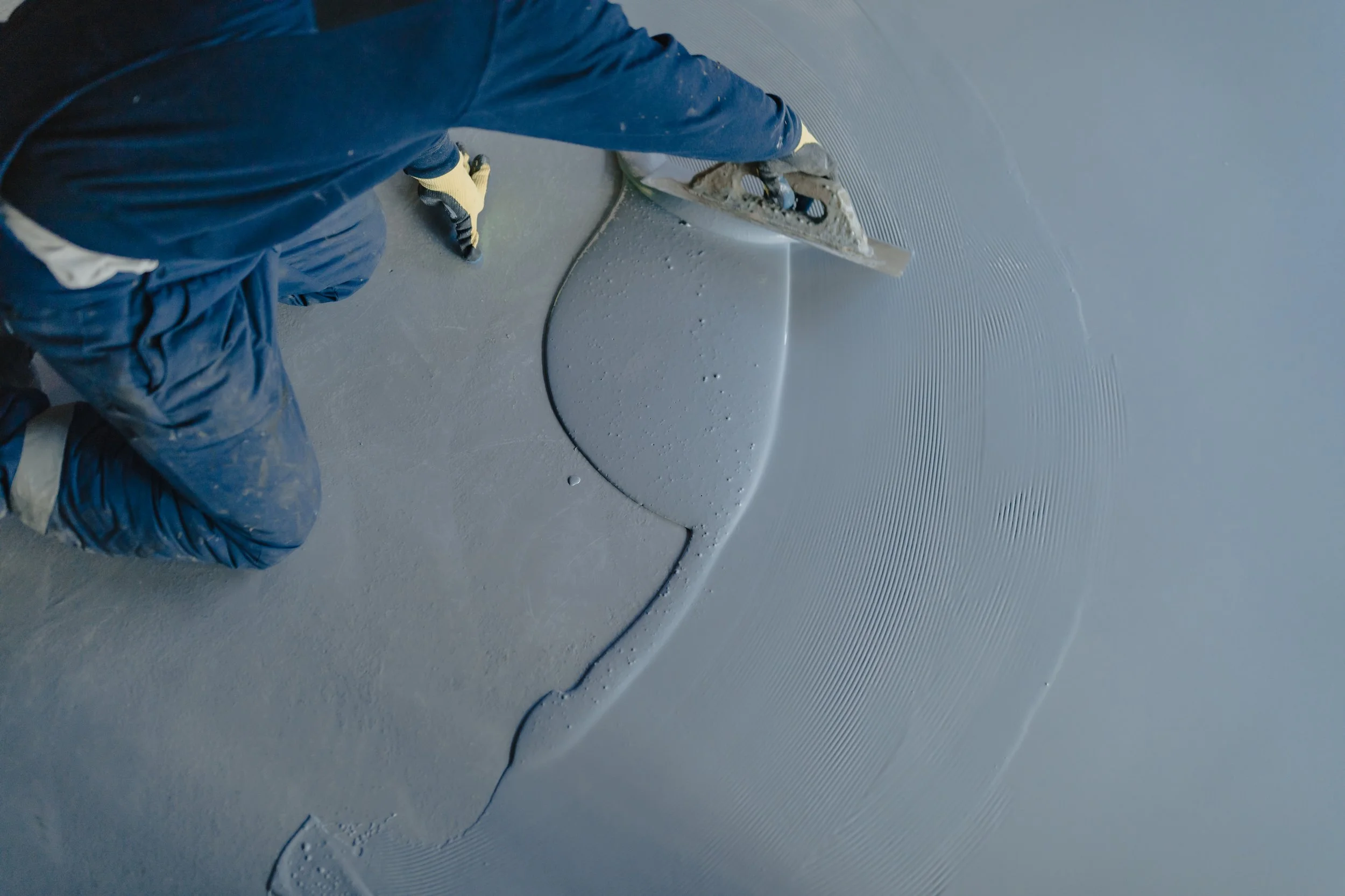 Worker smoothing wet cement on a floor with a trowel.