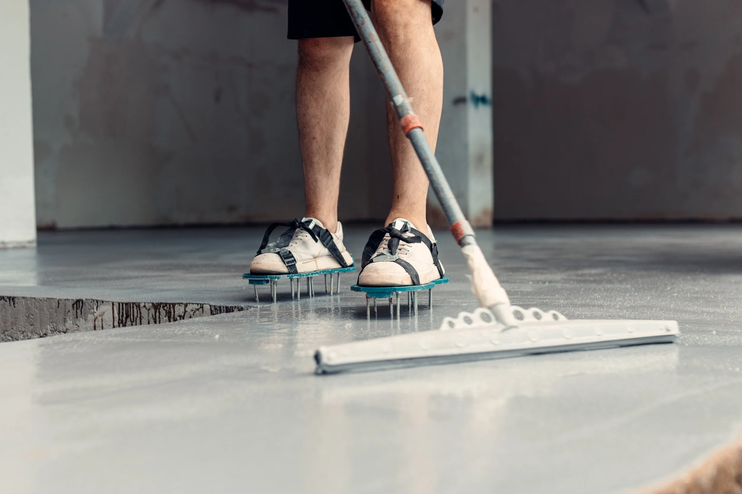 A person wearing athletic shoes on skate-like shoe attachments, cleaning or smoothing a concrete floor with a long-handled tool in an indoor space.