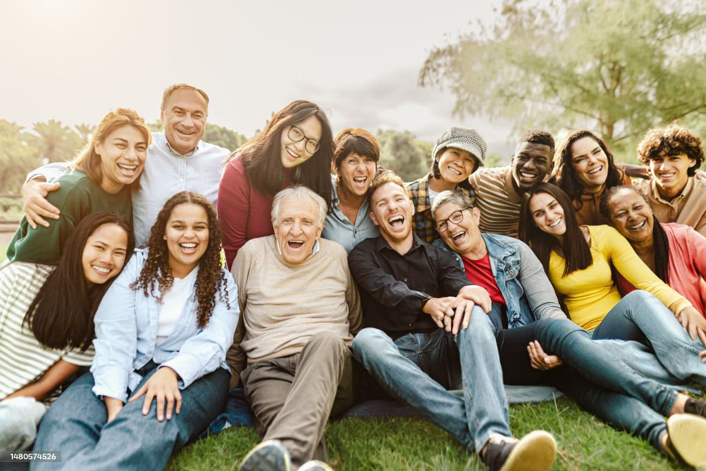 Group of 15 people sitting and standing on grass outdoors, smiling and laughing, in a park with trees.