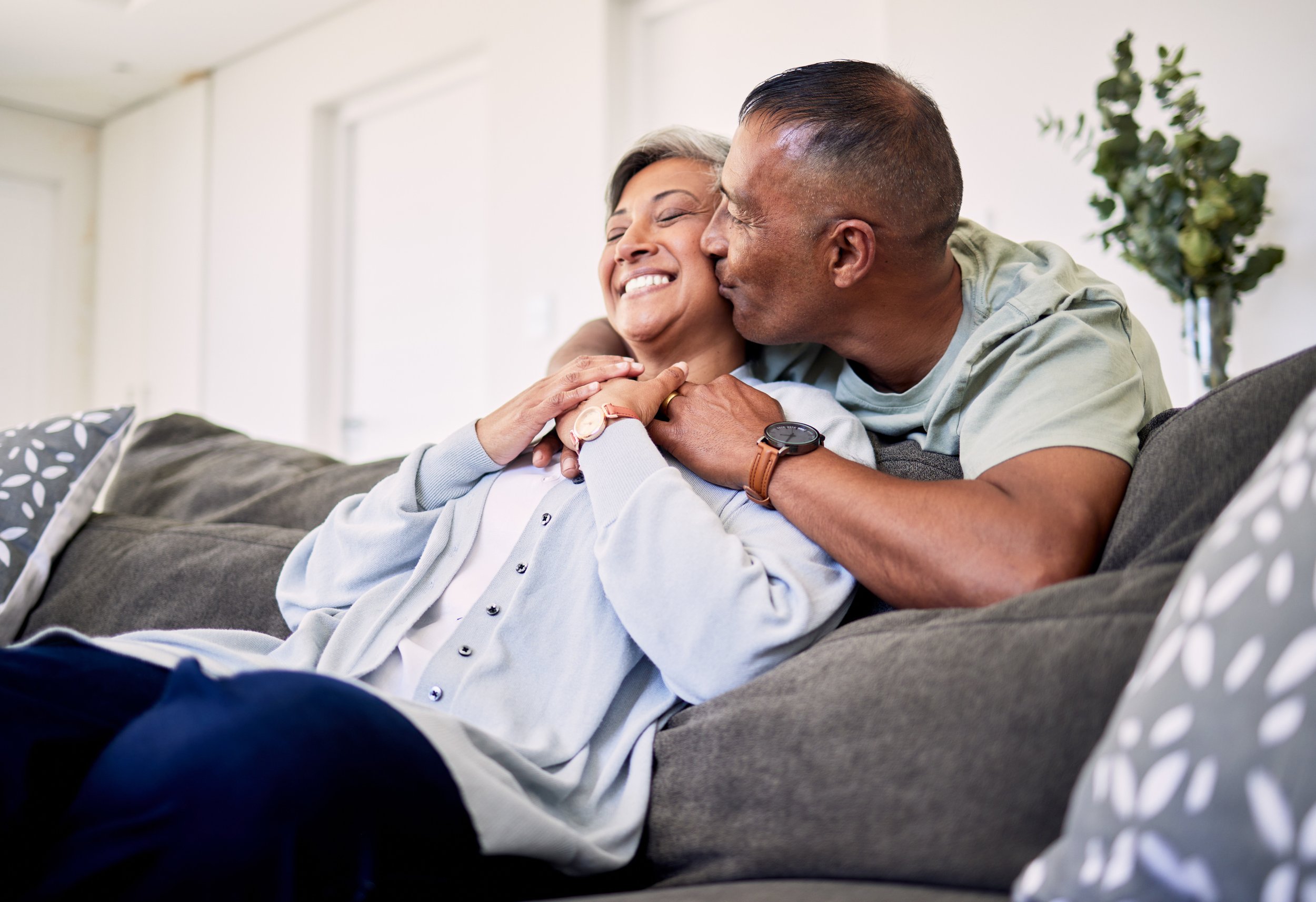 A man kisses an older woman on the cheek while sitting on a sofa, both smiling warmly, in a bright living room.