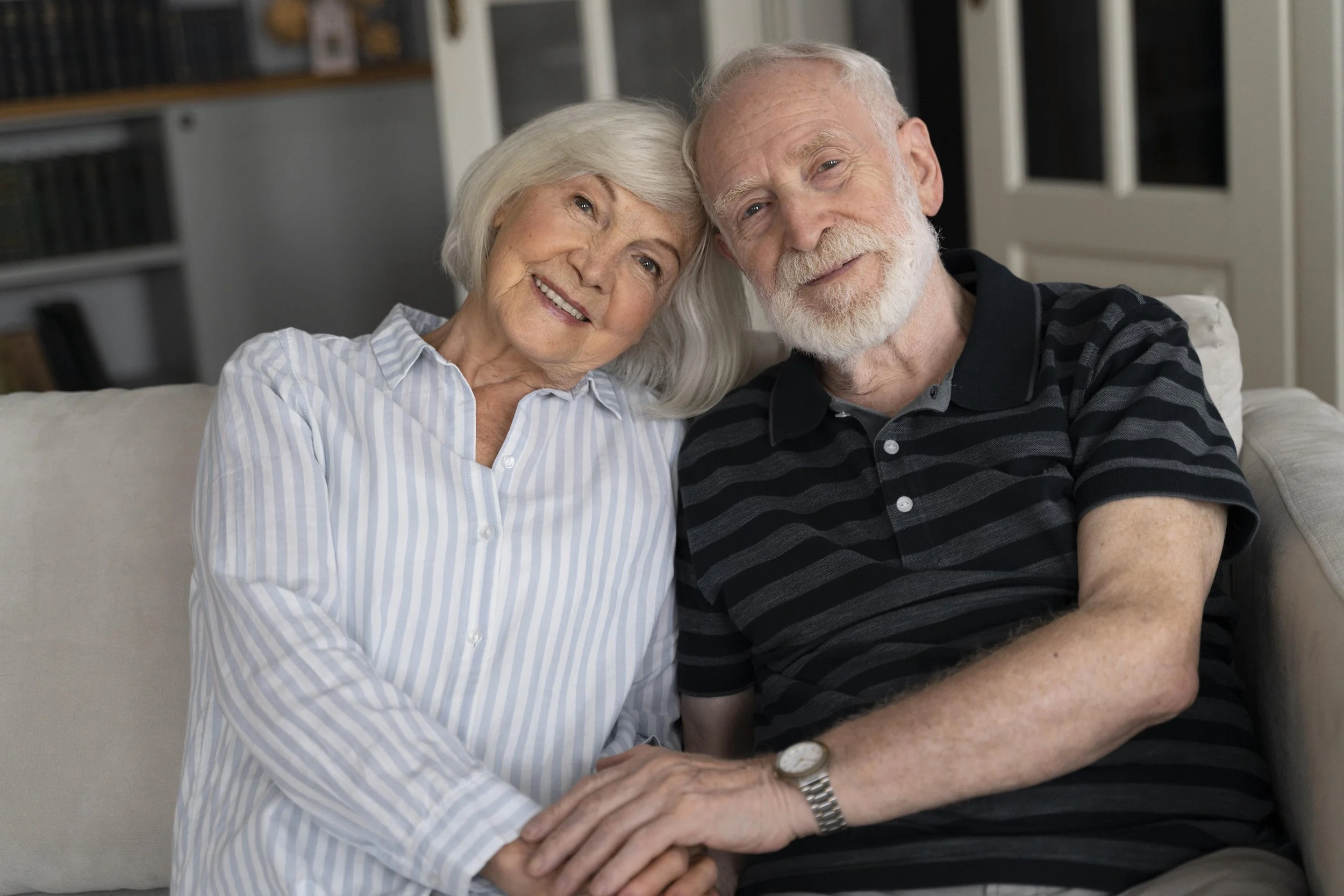 An elderly couple sitting on a couch, smiling and holding hands, in a cozy home setting.
