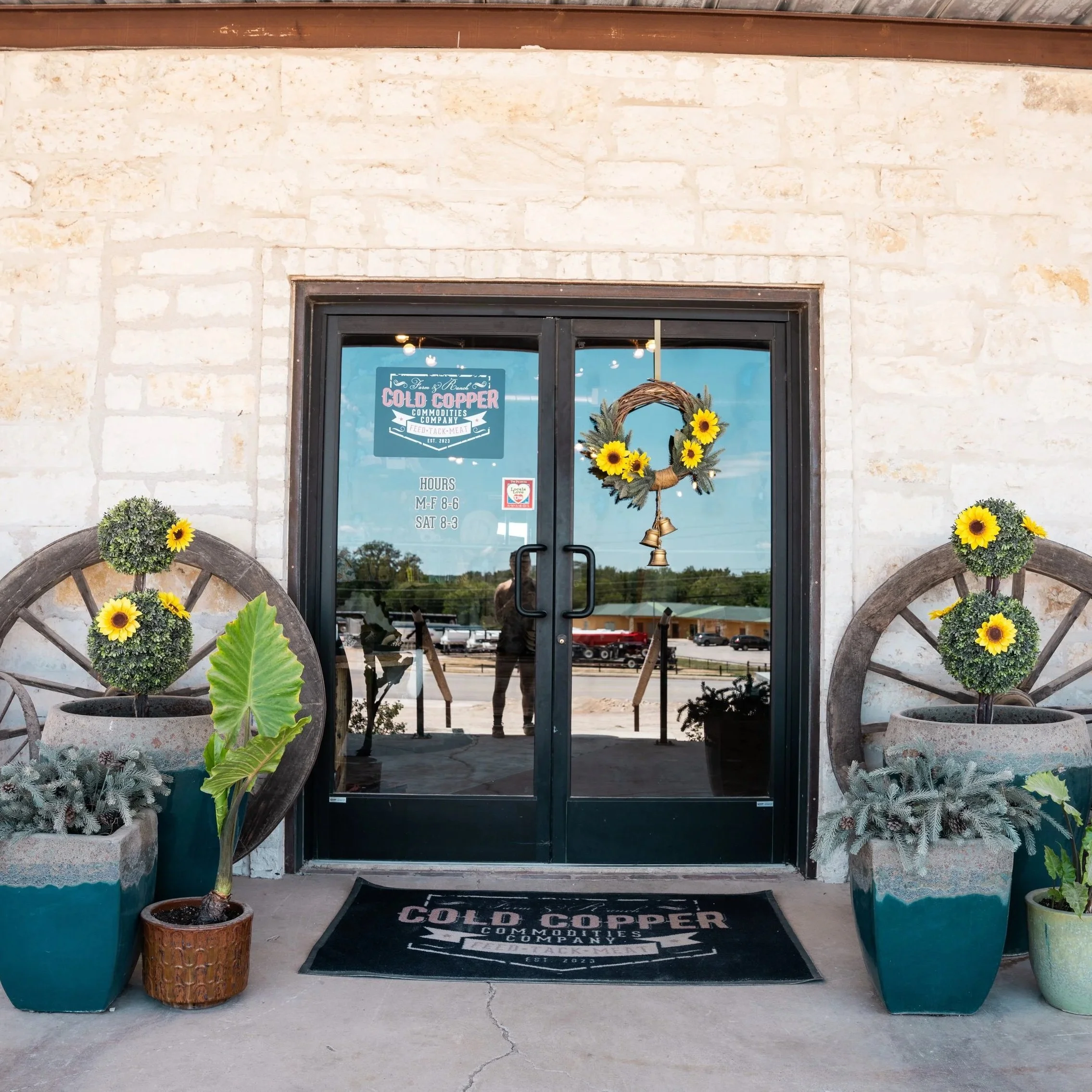 Burnet Feed Store entrance with glass door decorated with sunflower wreath, flanked by potted plants and sunflowers, with a black mat and a sign displaying business hours.