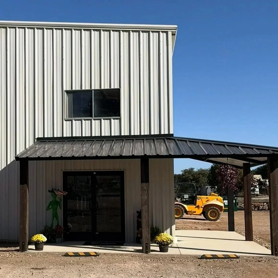 Marble Falls Feed store is A two-story building with metal siding, a small covered porch, and potted plants outside. A yellow construction vehicle is parked in the background under a clear blue sky.