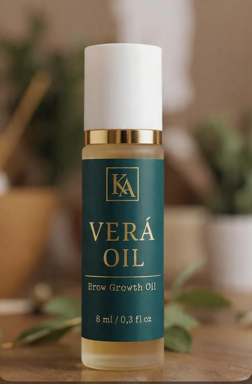 A small bottle of Vera Oil, labeled as brow growth oil, with a teal label, gold accents, and a white cap, sitting on a wooden surface with blurred plants in the background.