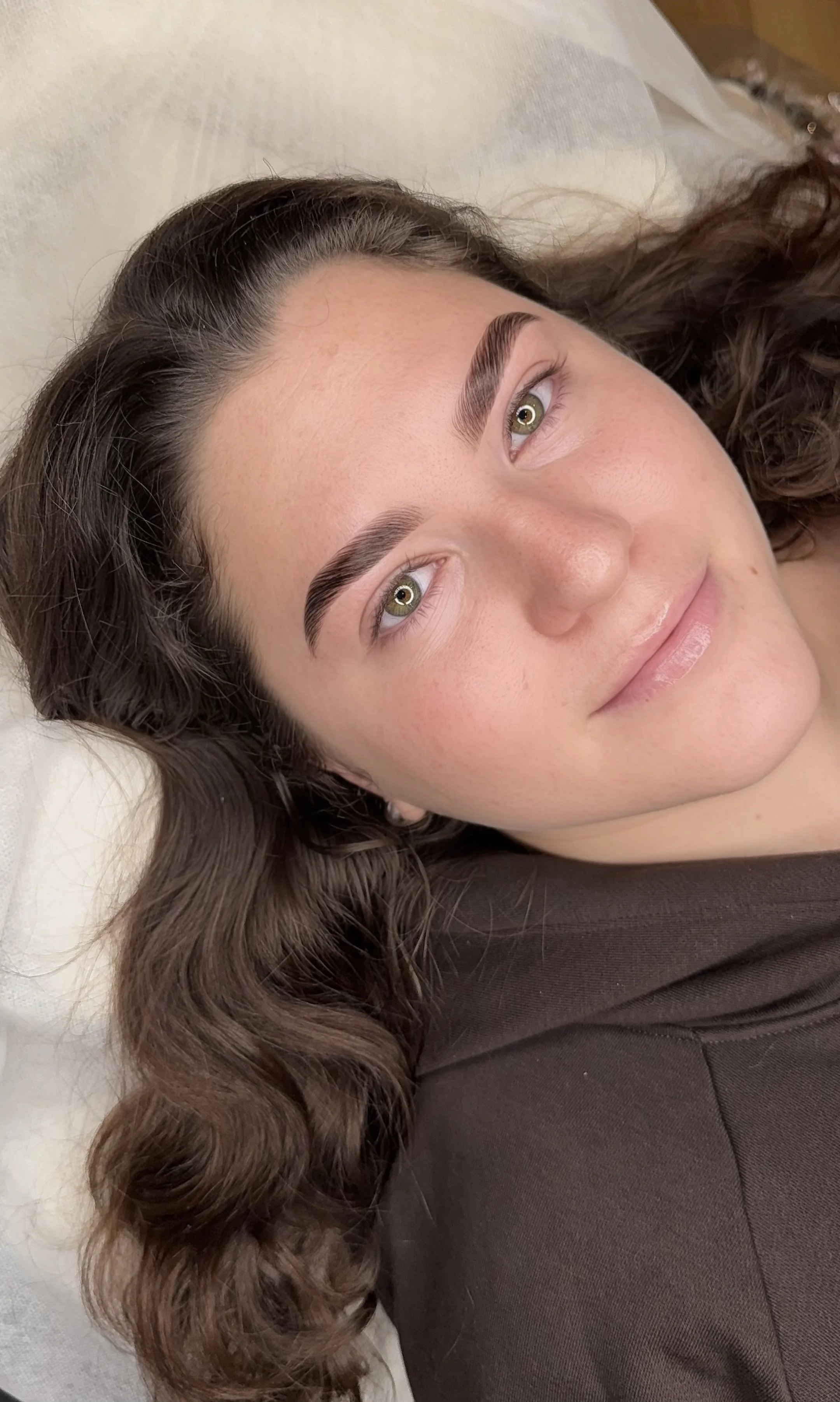 A young woman with wavy brown hair, fair skin, and green eyes lying on a white pillow, smiling gently at the camera, wearing a brown top.