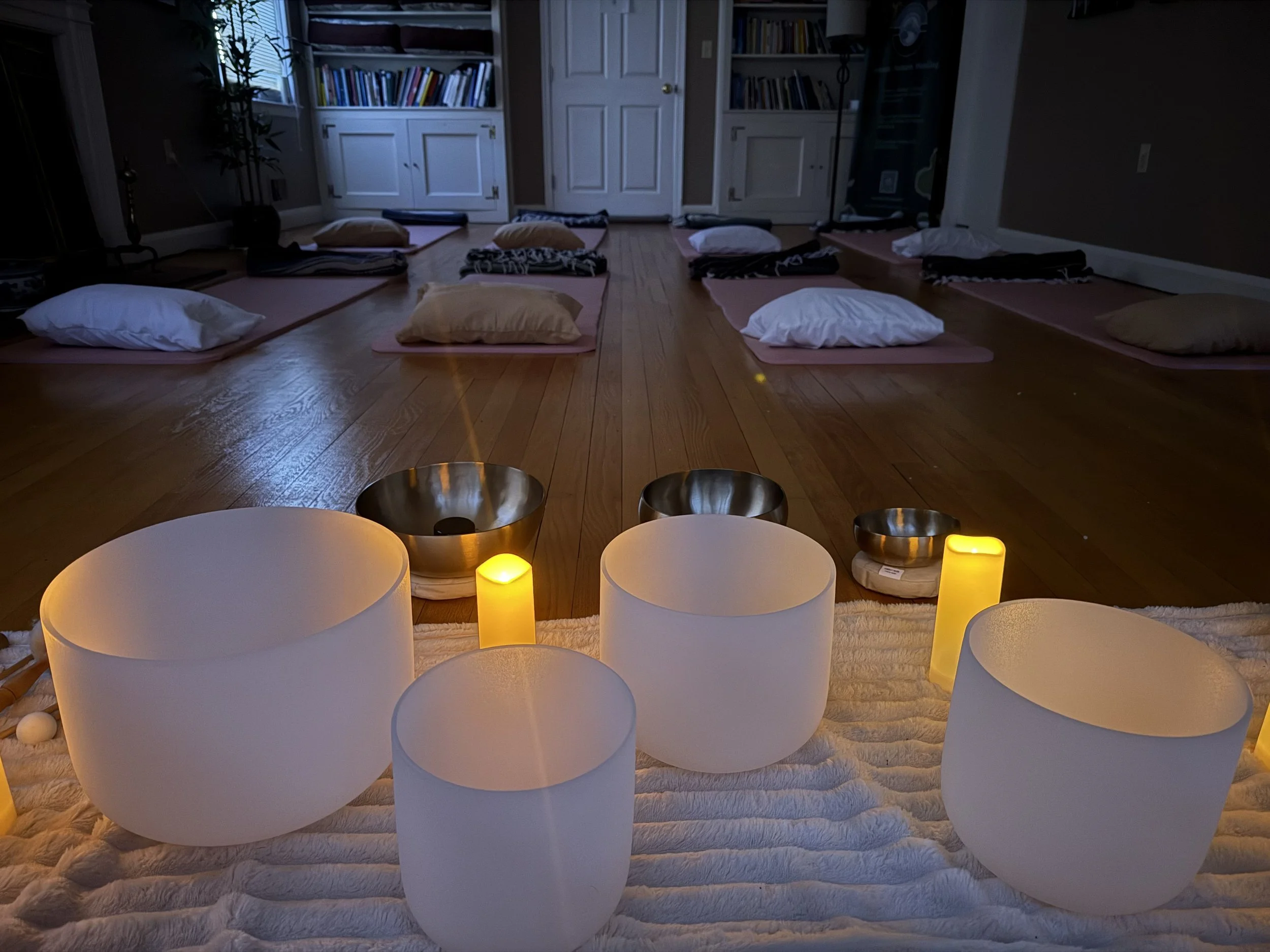 Indoor yoga or meditation space with stacked cushions, pillows, and blankets on a hardwood floor, illuminated by yellow candles, with bookshelves and closed doors in the background.