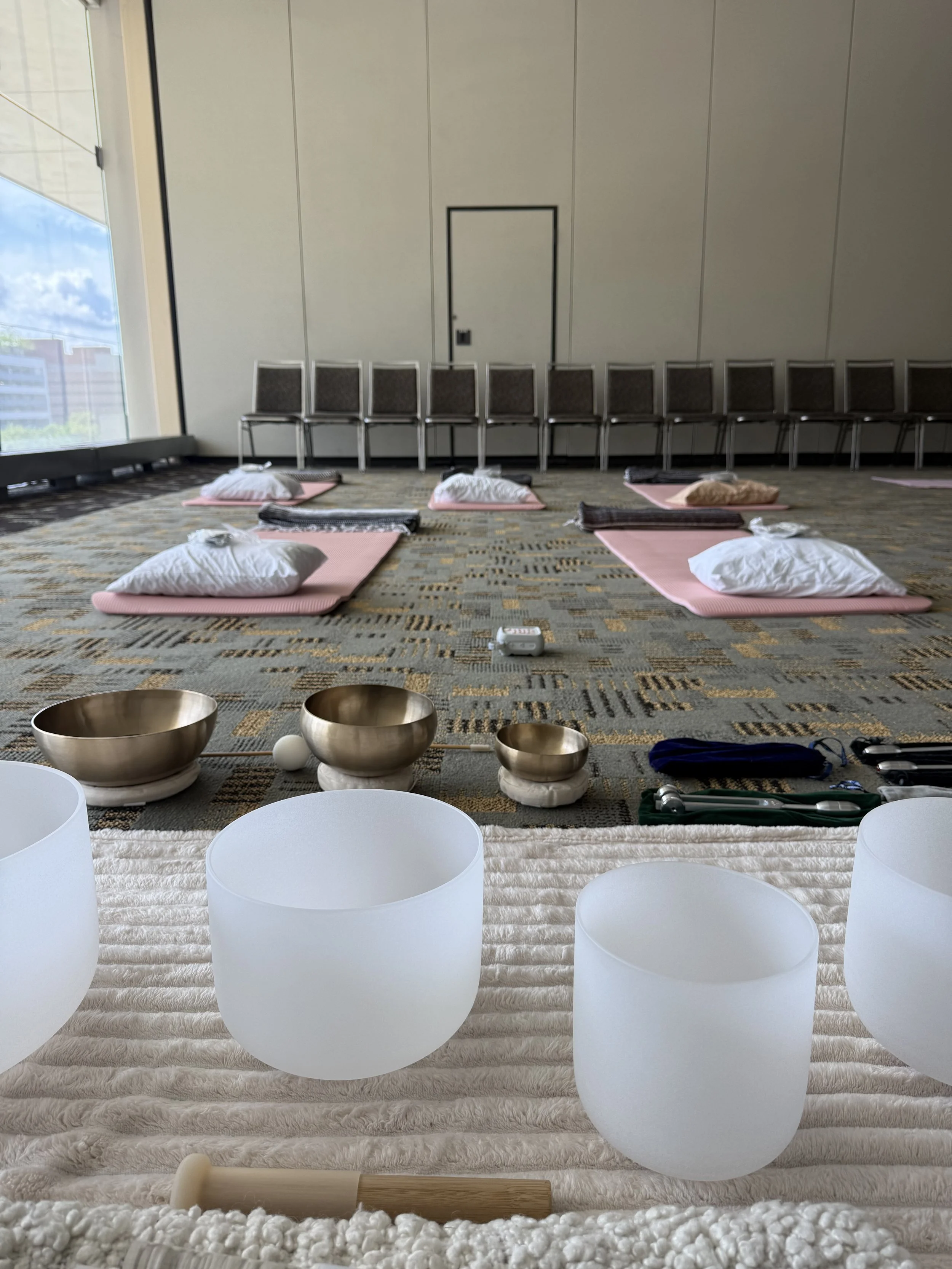 Yogurt bowl, Tibetan singing bowls, white chairs, meditation mats with pillows, and a drum on a beige textured rug in a spacious room with large windows and a city view.