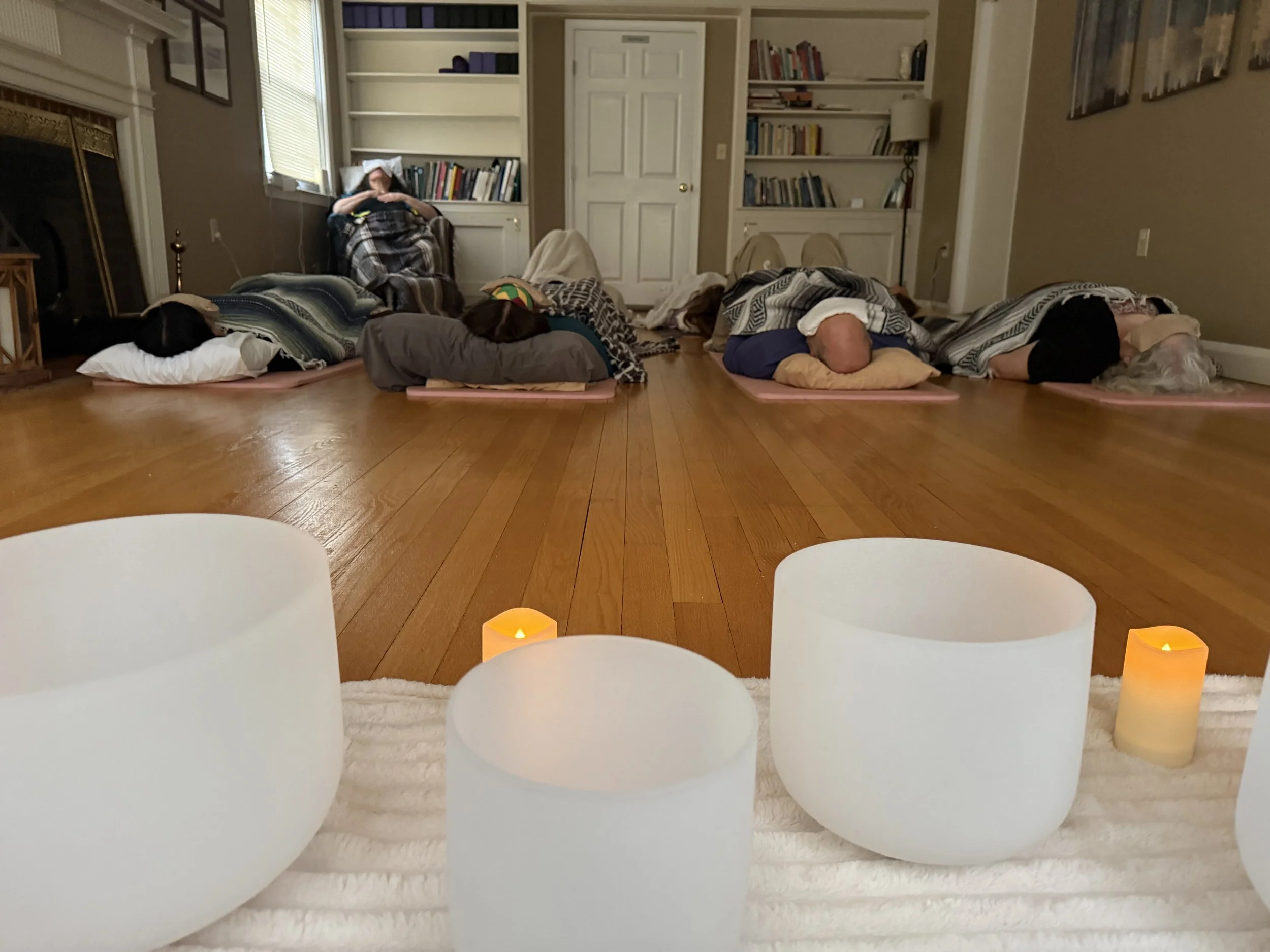 A group of people lying on yoga mats in a room, participating in a meditation or relaxation session, with candles and a cozy setting.