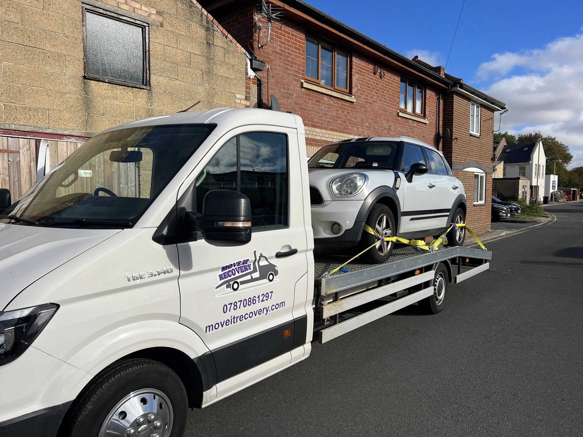 A white tow truck with a mini Cooper on its flatbed, parked on a residential street under a blue sky with some clouds. The truck has branding that says MOVE IT RECOVERY and provides a phone number and website.