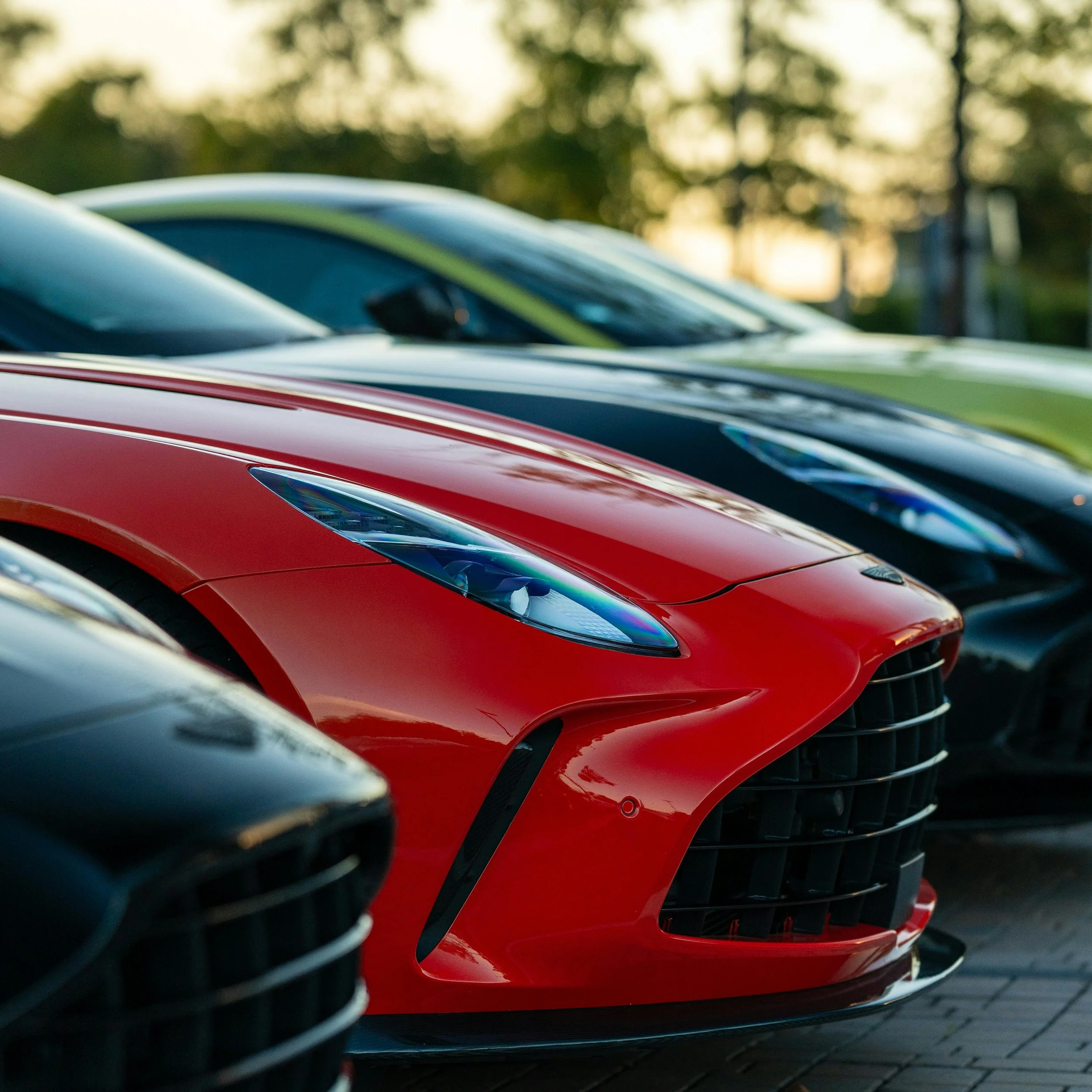 Close-up of a red sports car parked among other cars, with a sunset and trees in the background.