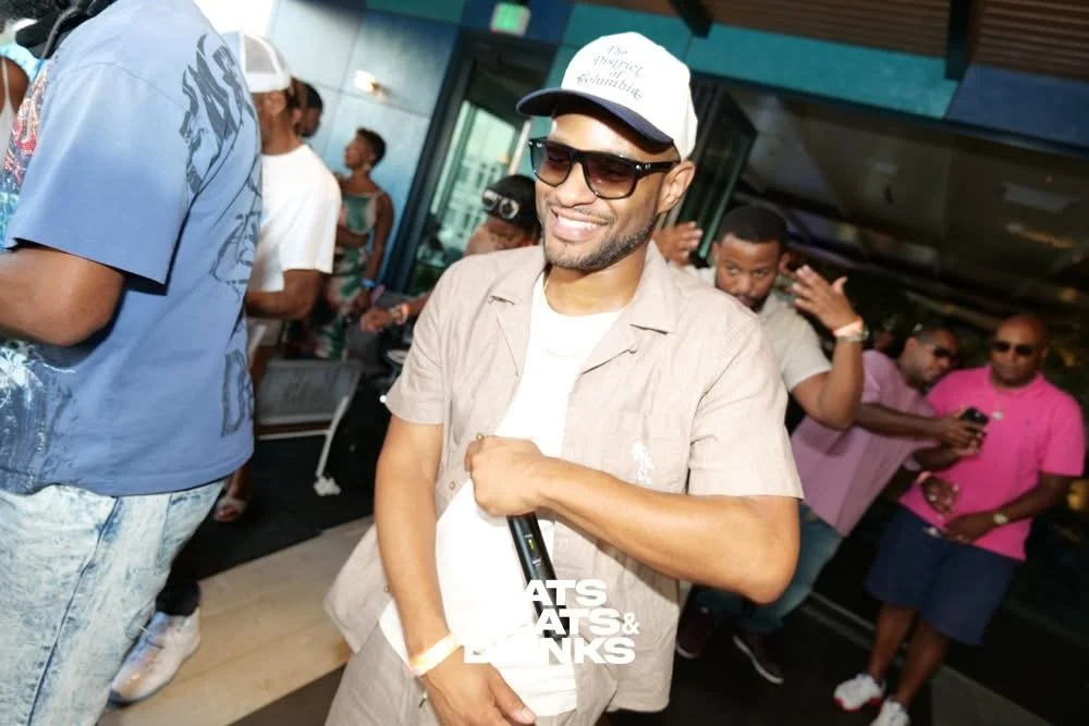 A man smiling and wearing sunglasses, a beige shirt, and a white baseball cap, holding a microphone at a party or social gathering.