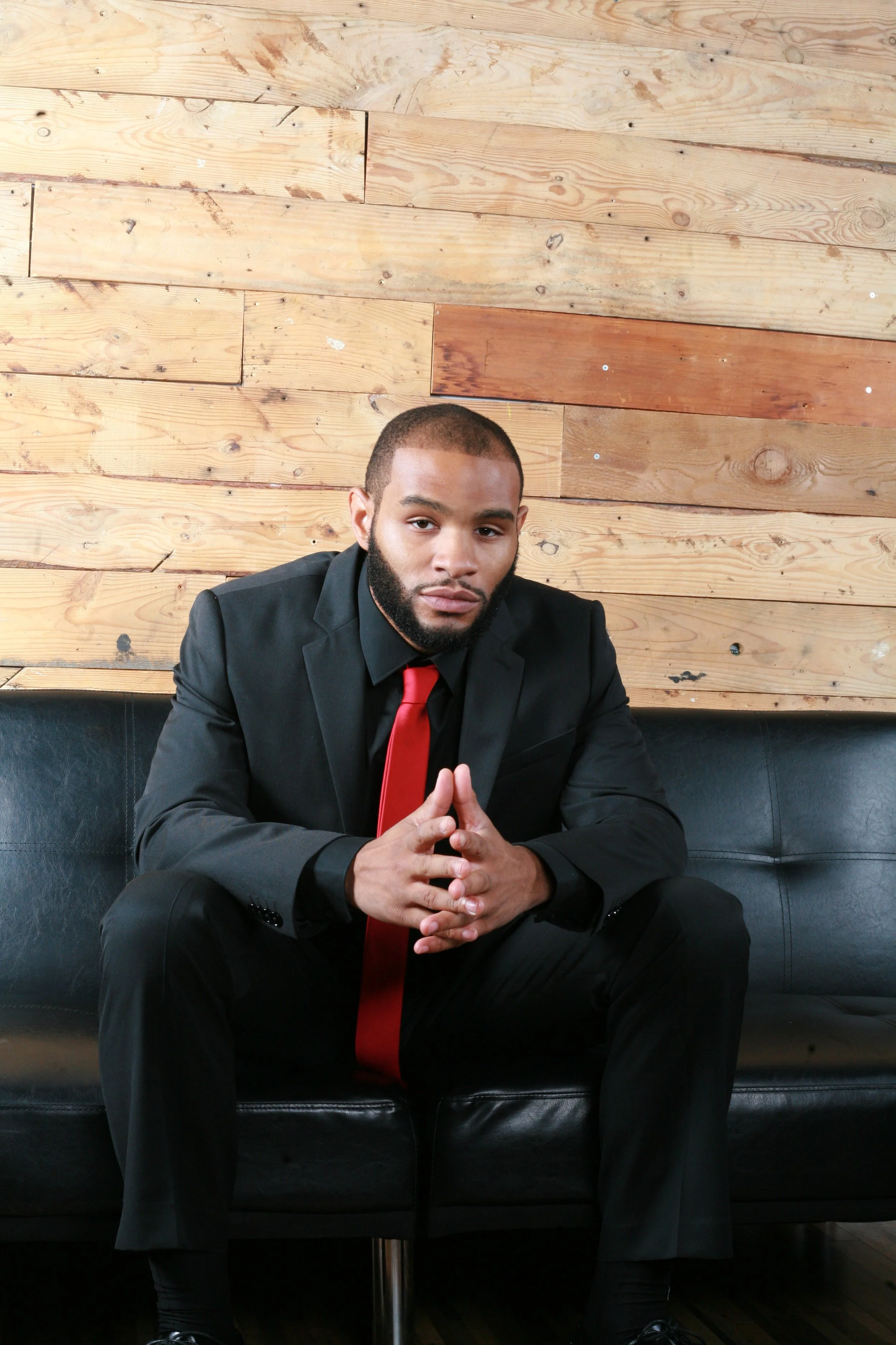 A man in a black suit with a red tie sitting on a black leather sofa against a wooden panel wall, with his hands clasped together.