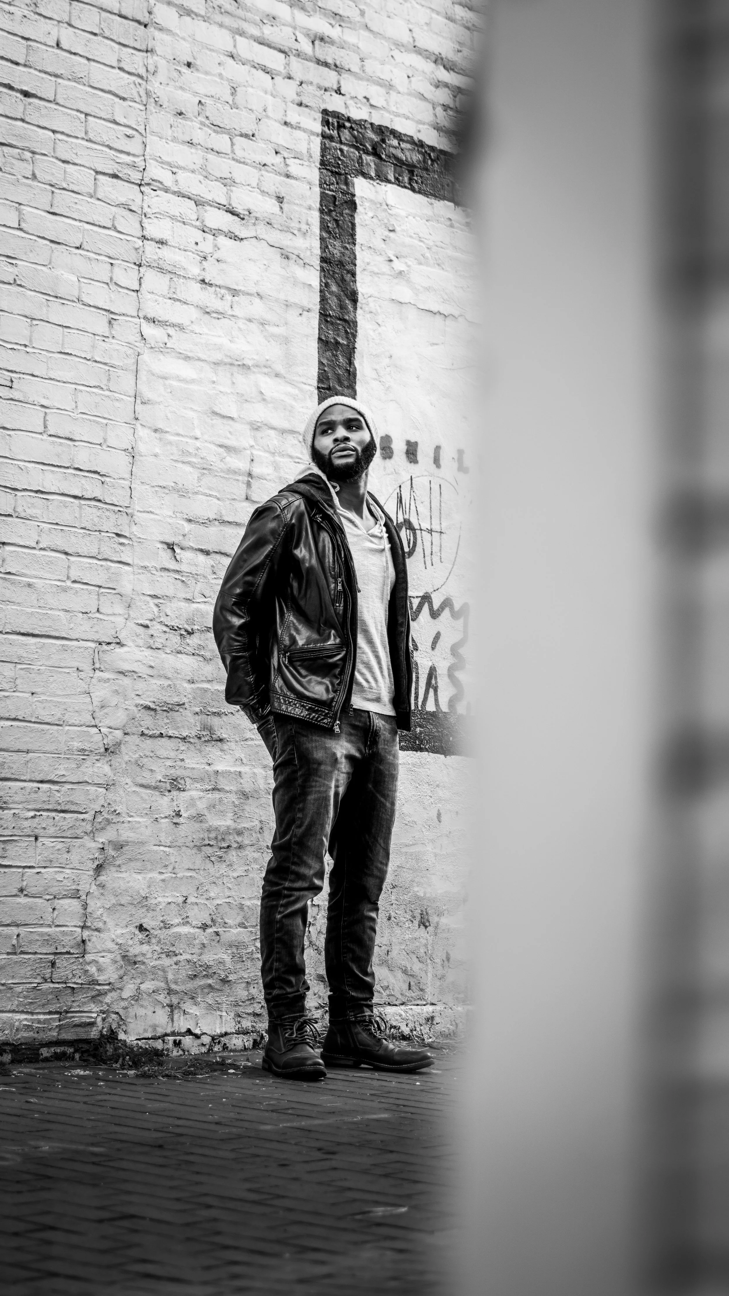 Black and white photo of DJ Reece standing against a brick wall, wearing a leather jacket, hoodie, jeans, and boots, with graffiti on the wall behind him.