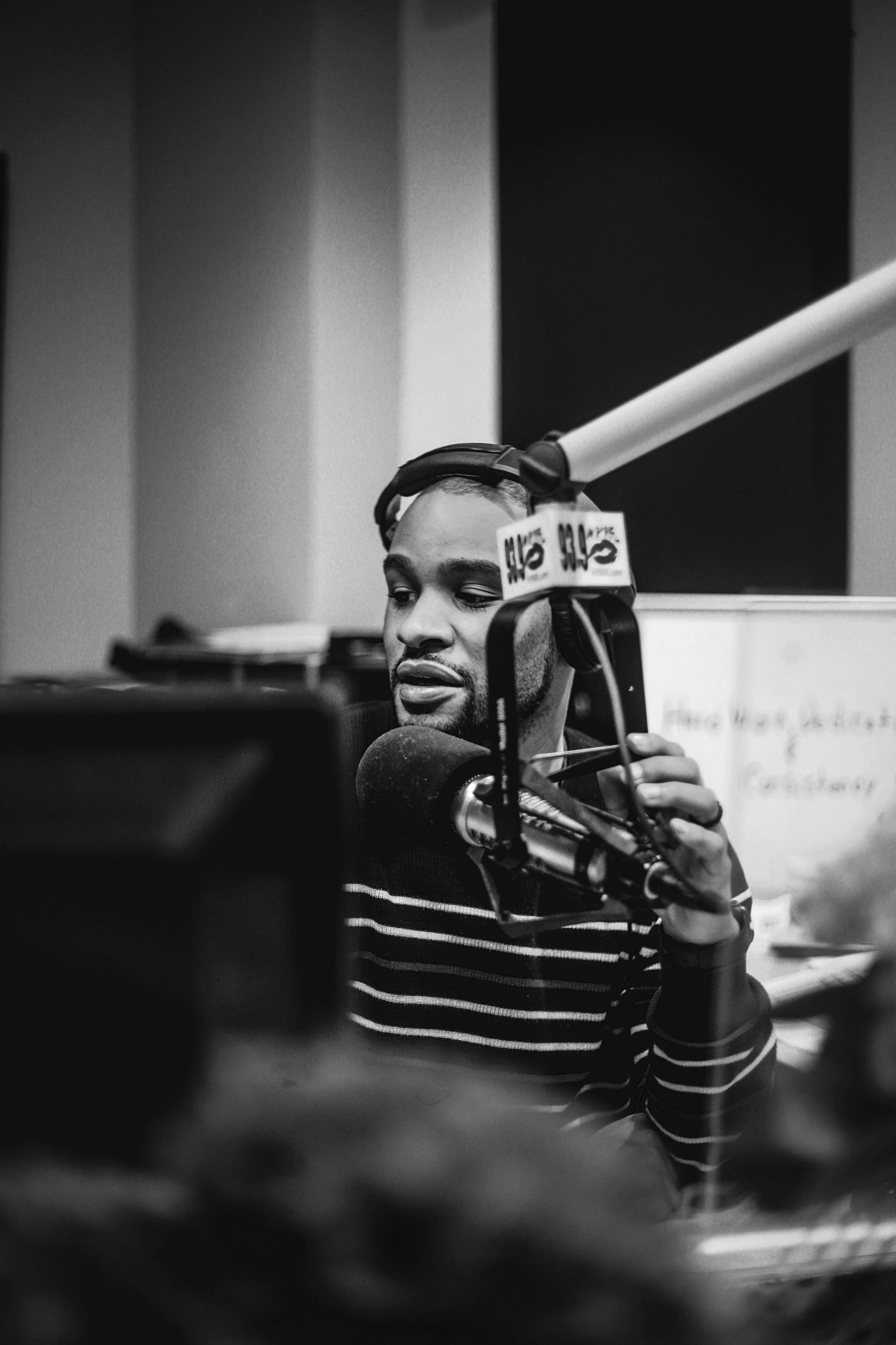 A man wearing headphones speaking into a microphone during a radio broadcast or recording session, with a window and some papers in the background.