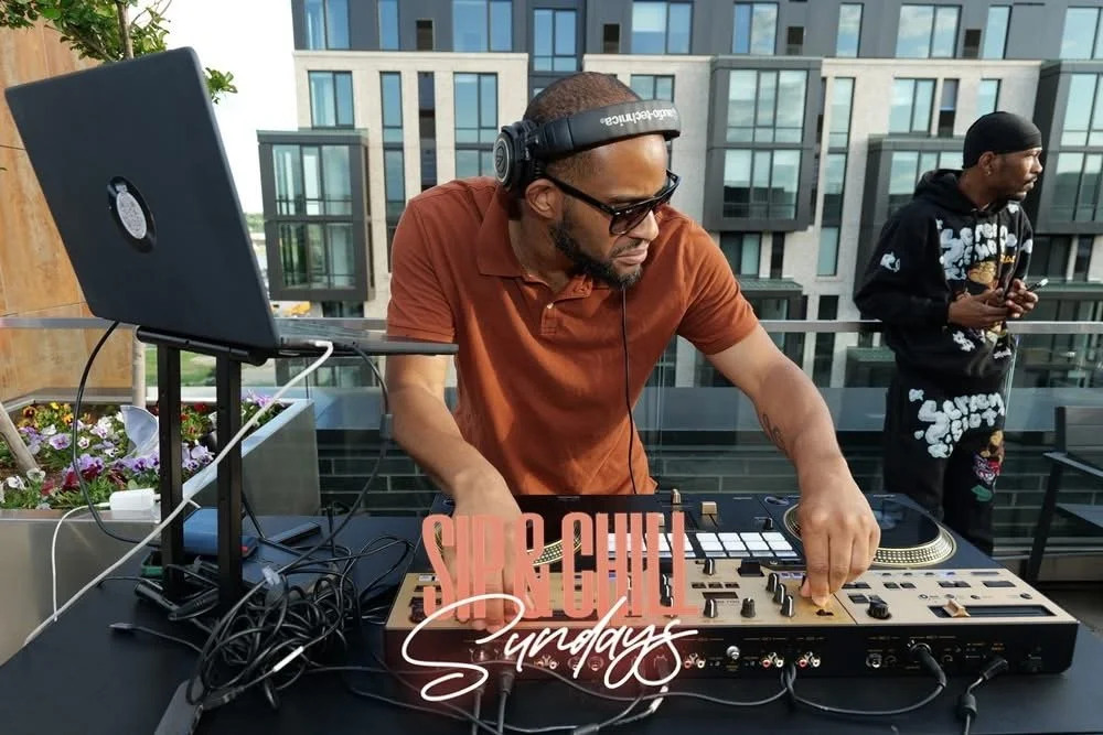 DJ wearing sunglasses and headset, mixing music on a DJ controller, with a laptop and cables nearby, outdoors on a rooftop with a cityscape background, and another person in the background holding a phone.
