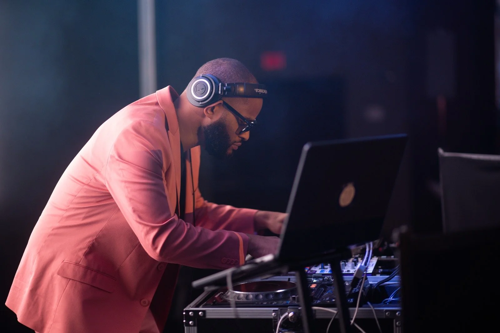 A DJ wearing a pink blazer, headphones, and glasses is working at a DJ booth with a laptop and audio equipment in a dark room.