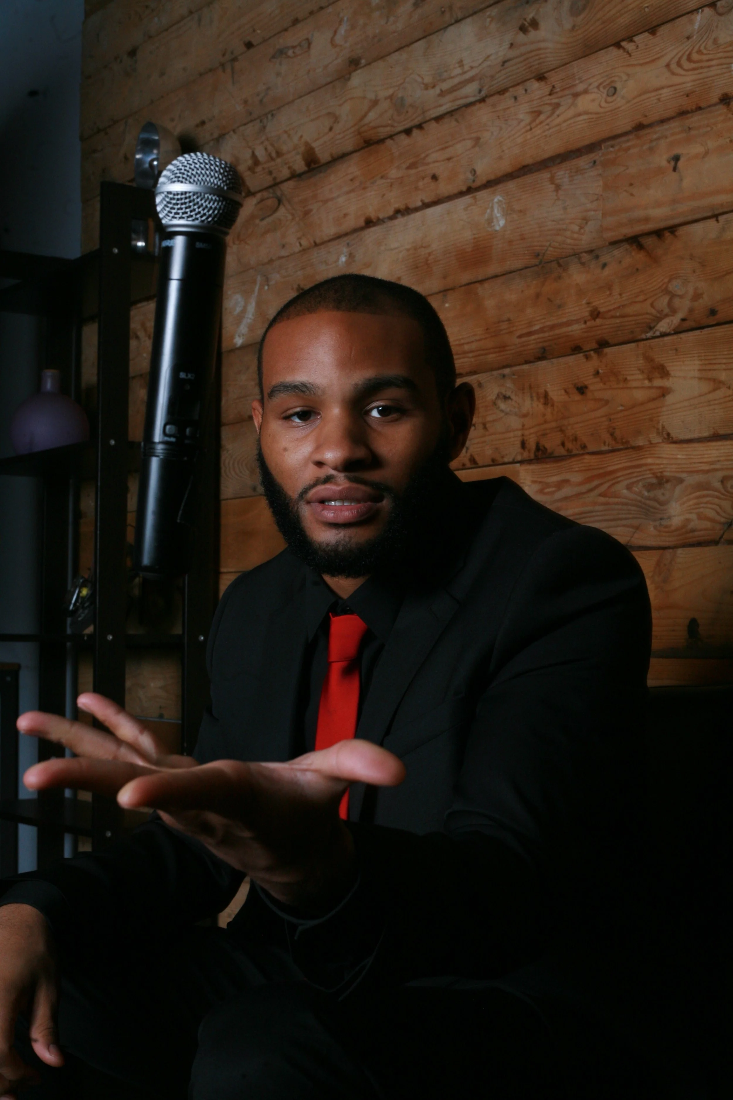 A man with a beard in a black suit and red tie sitting indoors against a wooden wall, with a microphone nearby.