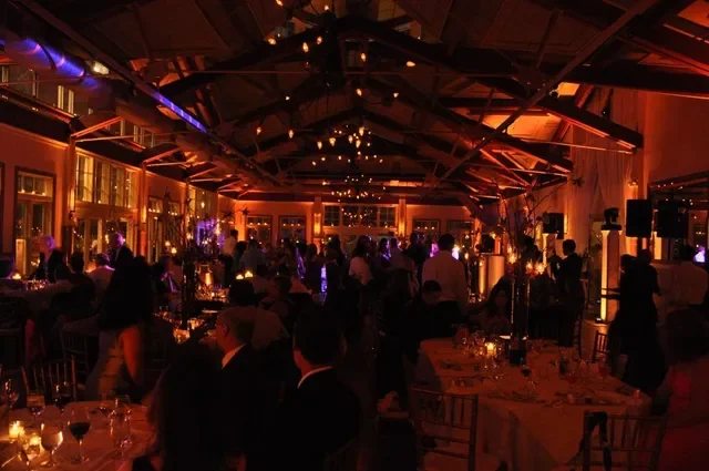 People gathered in a warmly lit wooden banquet hall with high ceilings and uplights provided by DJ Reece