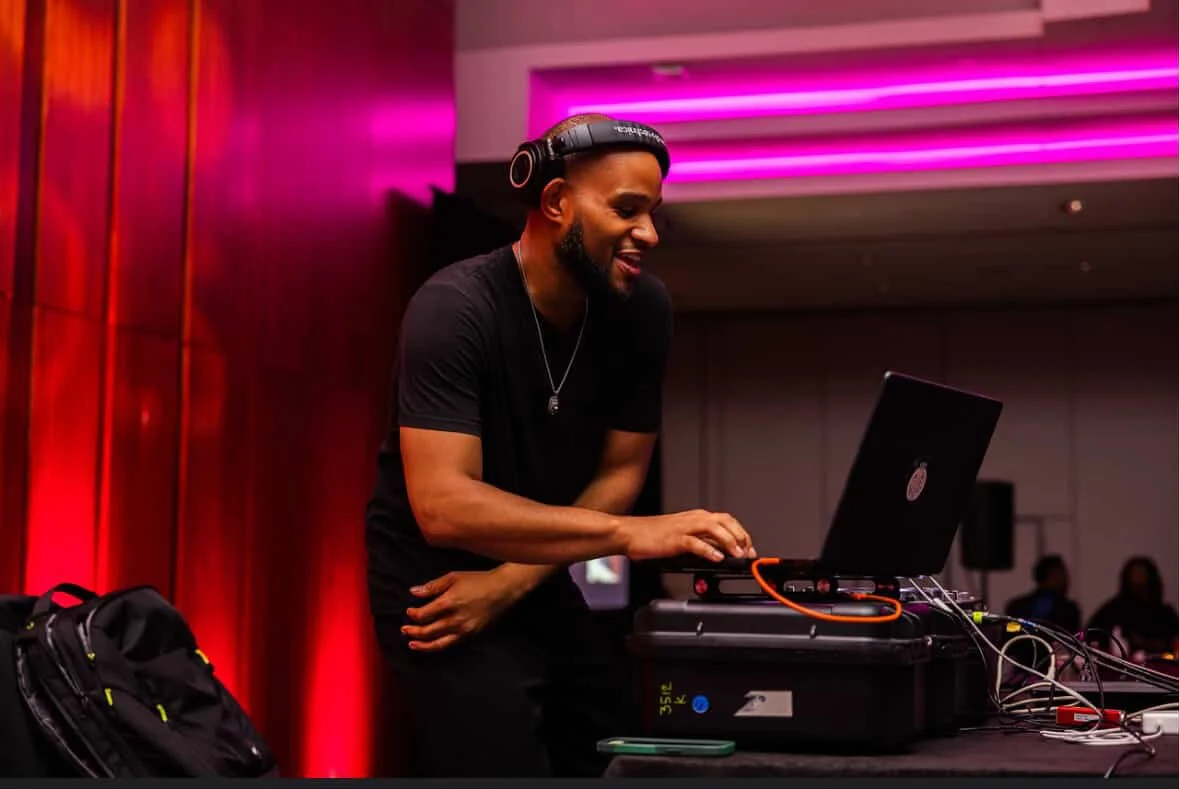A DJ wearing headphones and a black t-shirt in a nightclub or party setting, using laptop and DJ equipment with pink neon lights overhead.