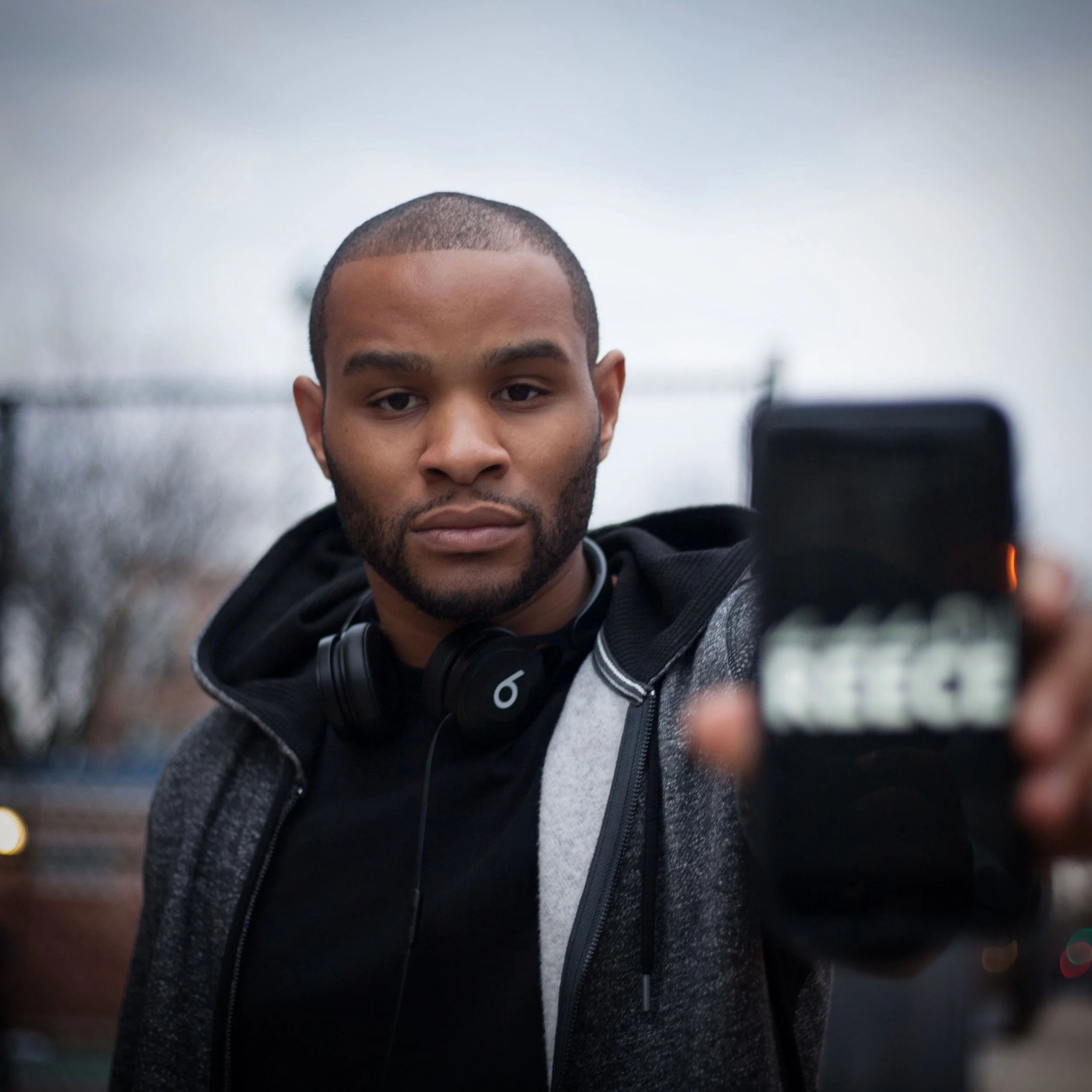 A young man with a beard and short hair taking a selfie outdoors in a cloudy environment, wearing headphones around his neck and a casual hoodie.