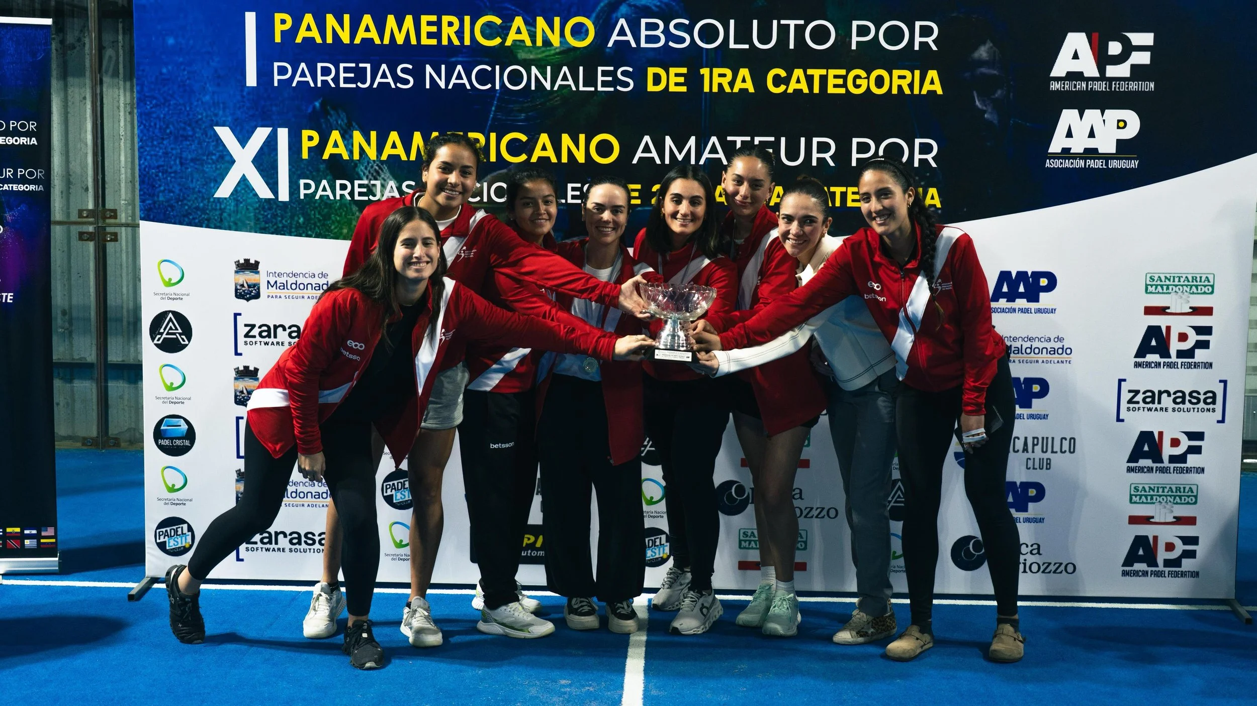 Un grupo de jugadoras de pádel en uniforme rojo y blanco, celebrando con un trofeo en una cancha de pádel, durante un evento de pádel en Uruguay.