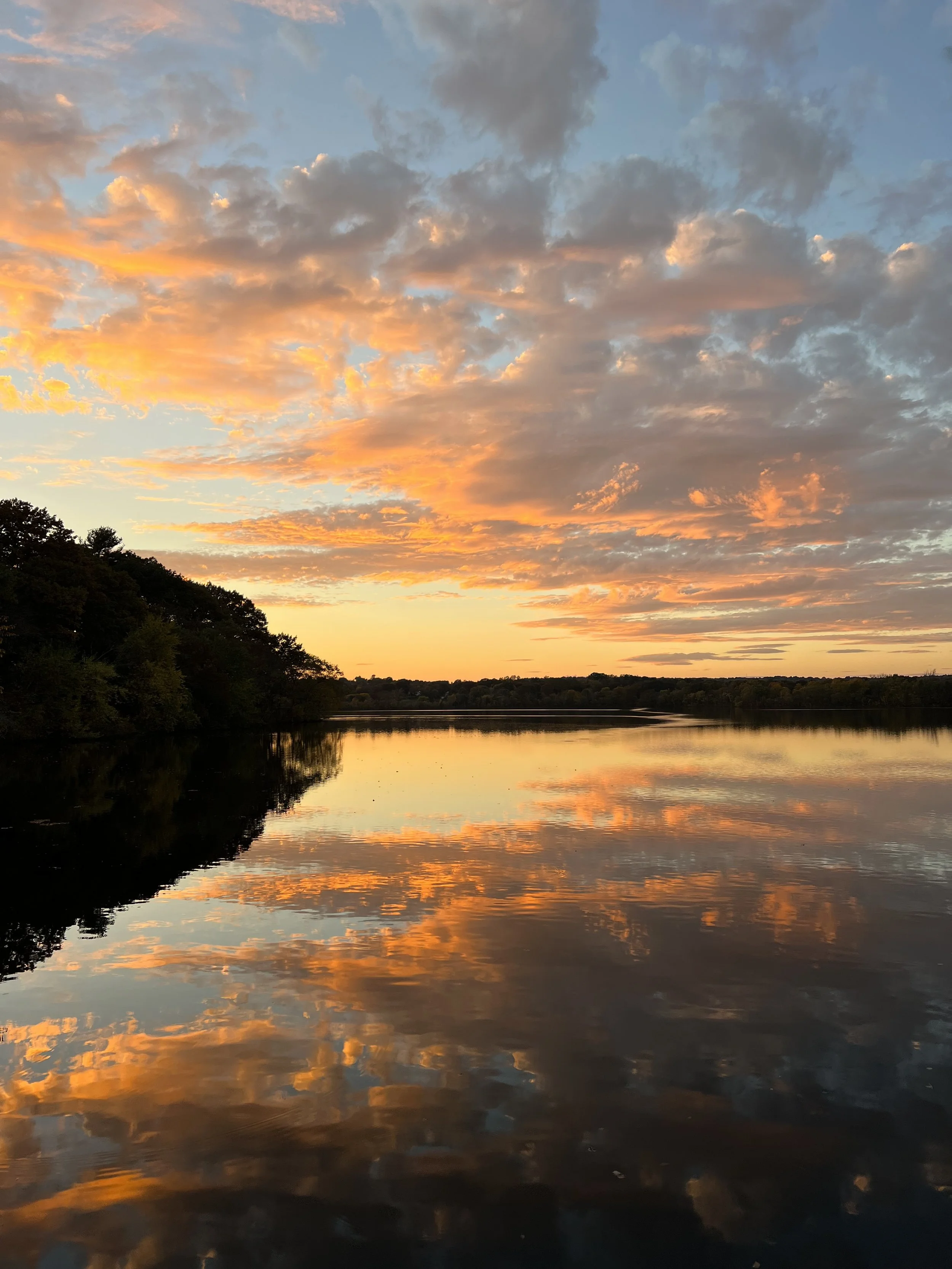 Sunset over a calm lake with colorful clouds and trees on the shoreline reflected in the water.
