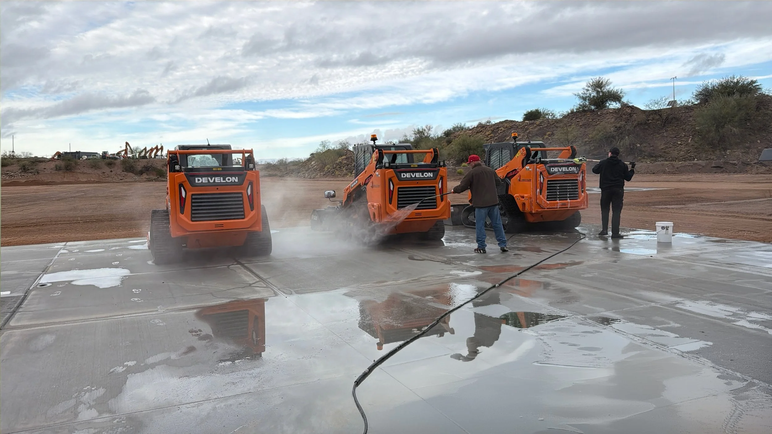 Two orange DeLEON industrial machines working outdoors on a concrete surface, with two men operating and cleaning it, while dirt and equipment are visible in the background under a partly cloudy sky.