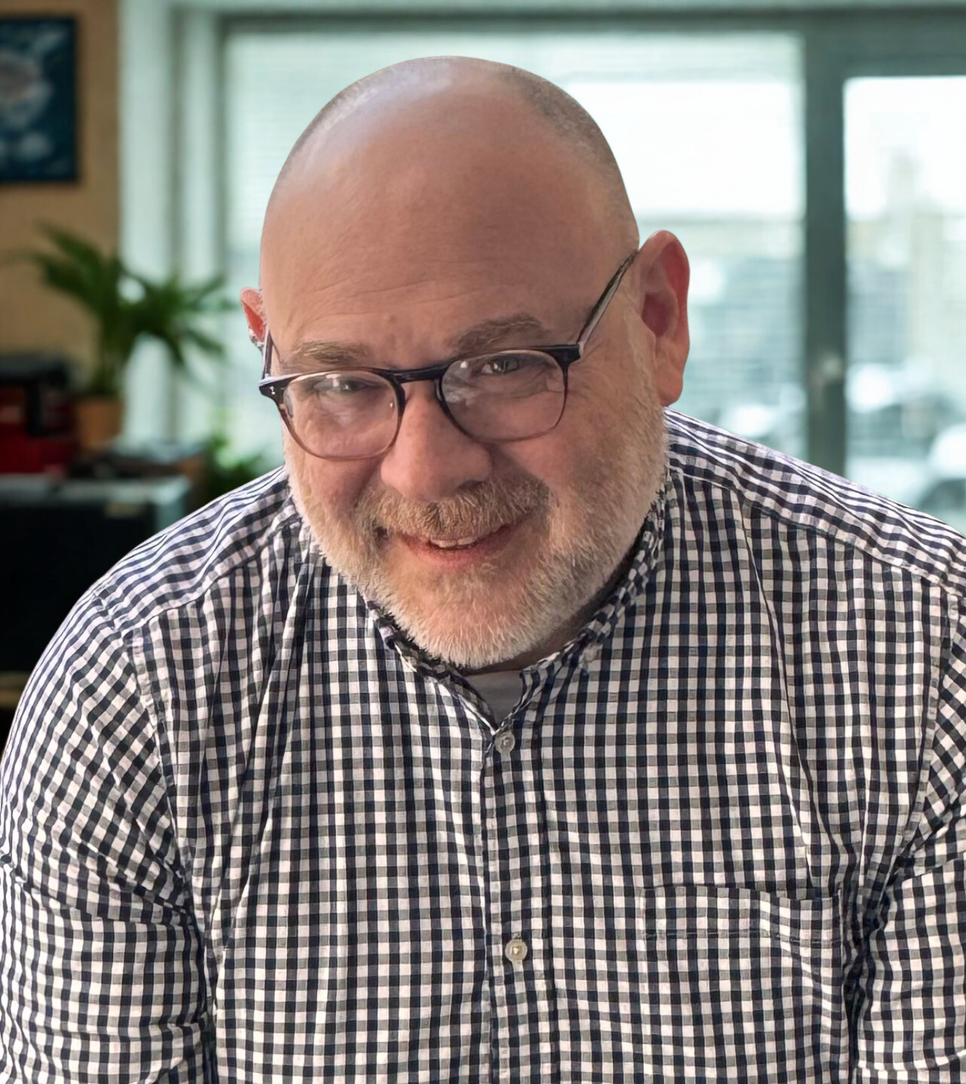 A smiling, mature man with glasses and a gray beard, wearing a checkered shirt, inside an ofice with large windows, a potted plant, and some office furniture.