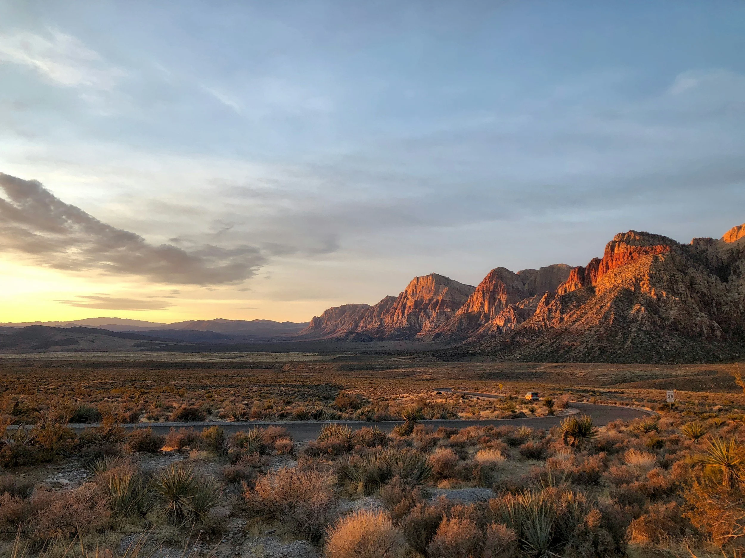 Nevada desert landscape at sunset with mountains in the background, sparse bushes, and a winding road in the foreground.