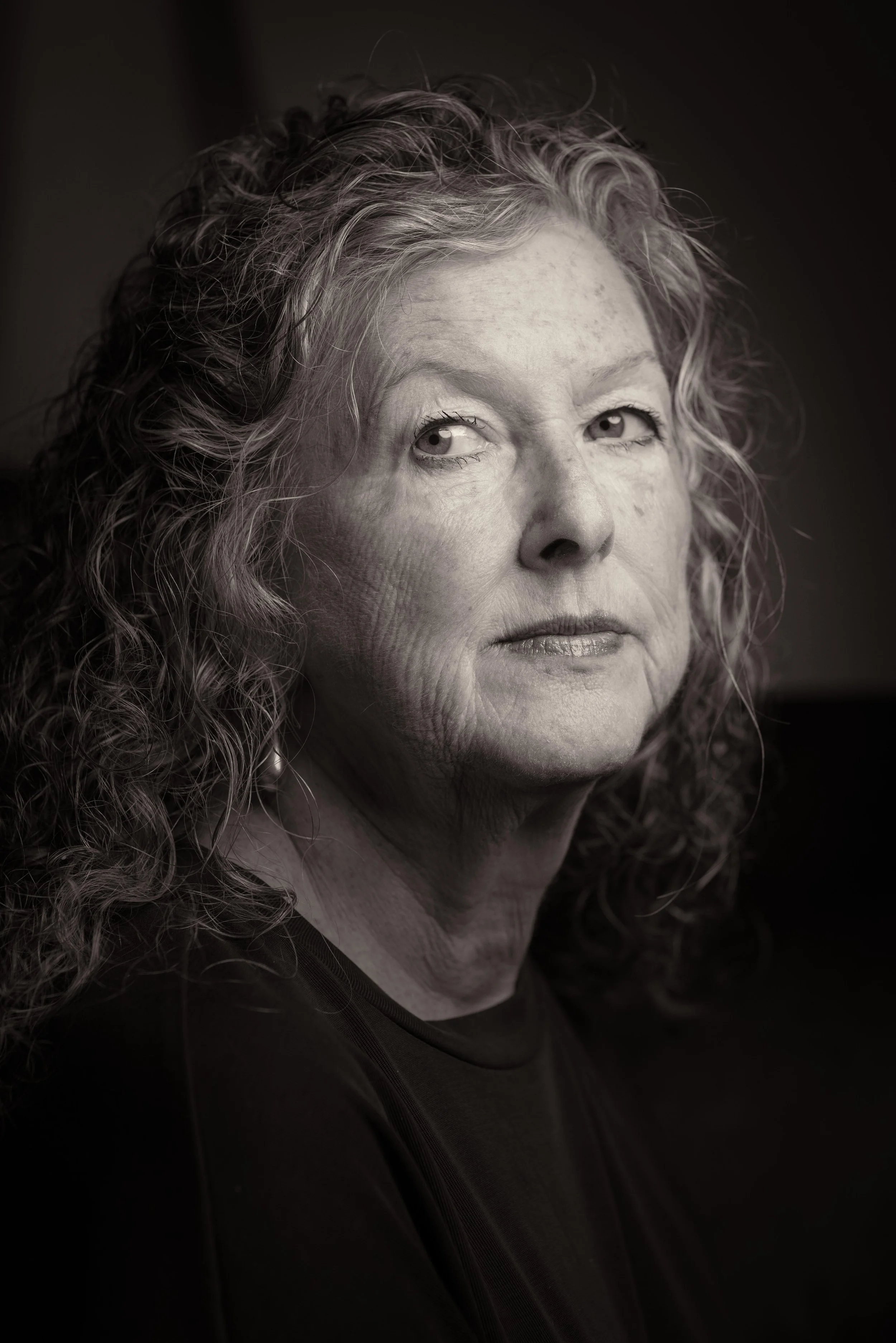 Close-up black-and-white portrait of author, Kathleen Bethel, with curly hair looking slightly to the right, wearing a dark top.