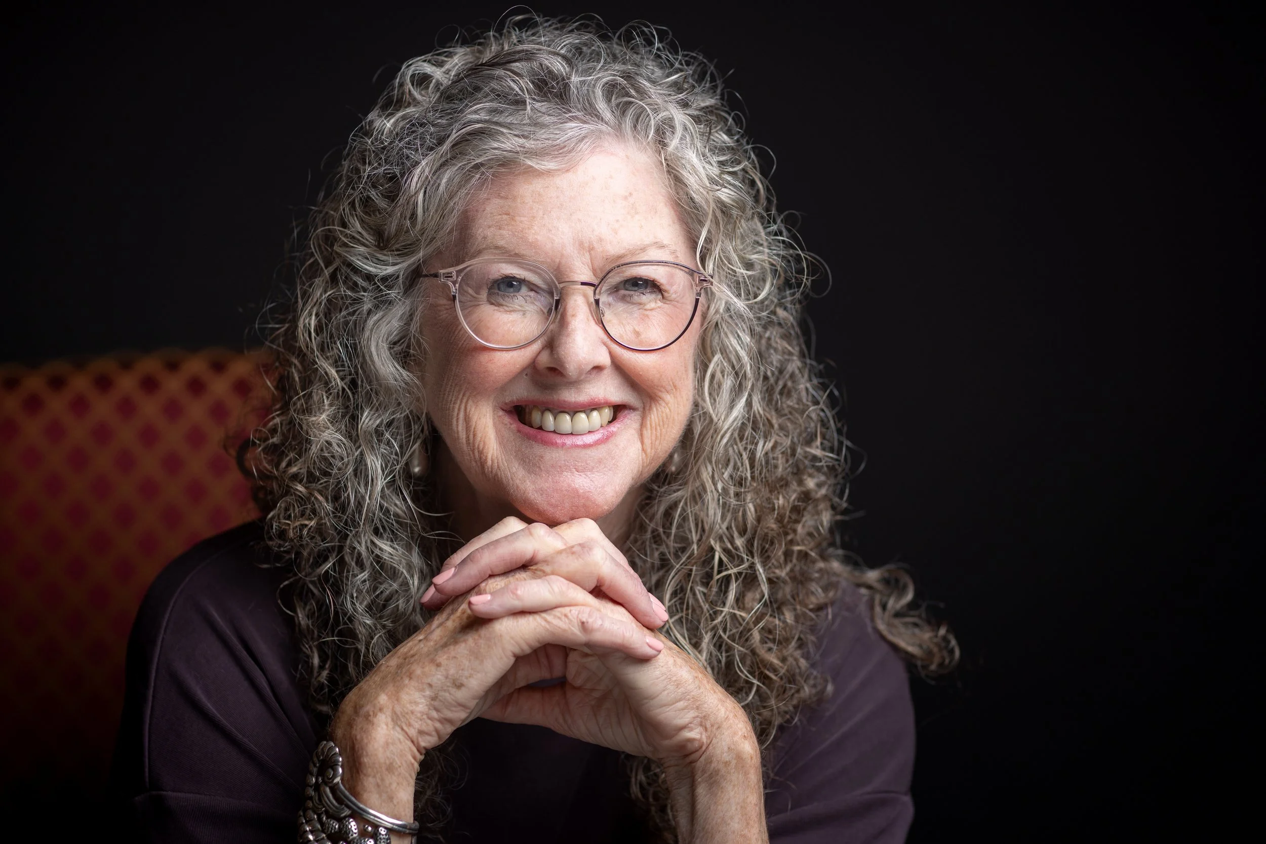 Portrait of author, Kathleen Bethel,  with curly gray hair, wearing glasses, smiling, and resting her chin on her hands, against a dark background.