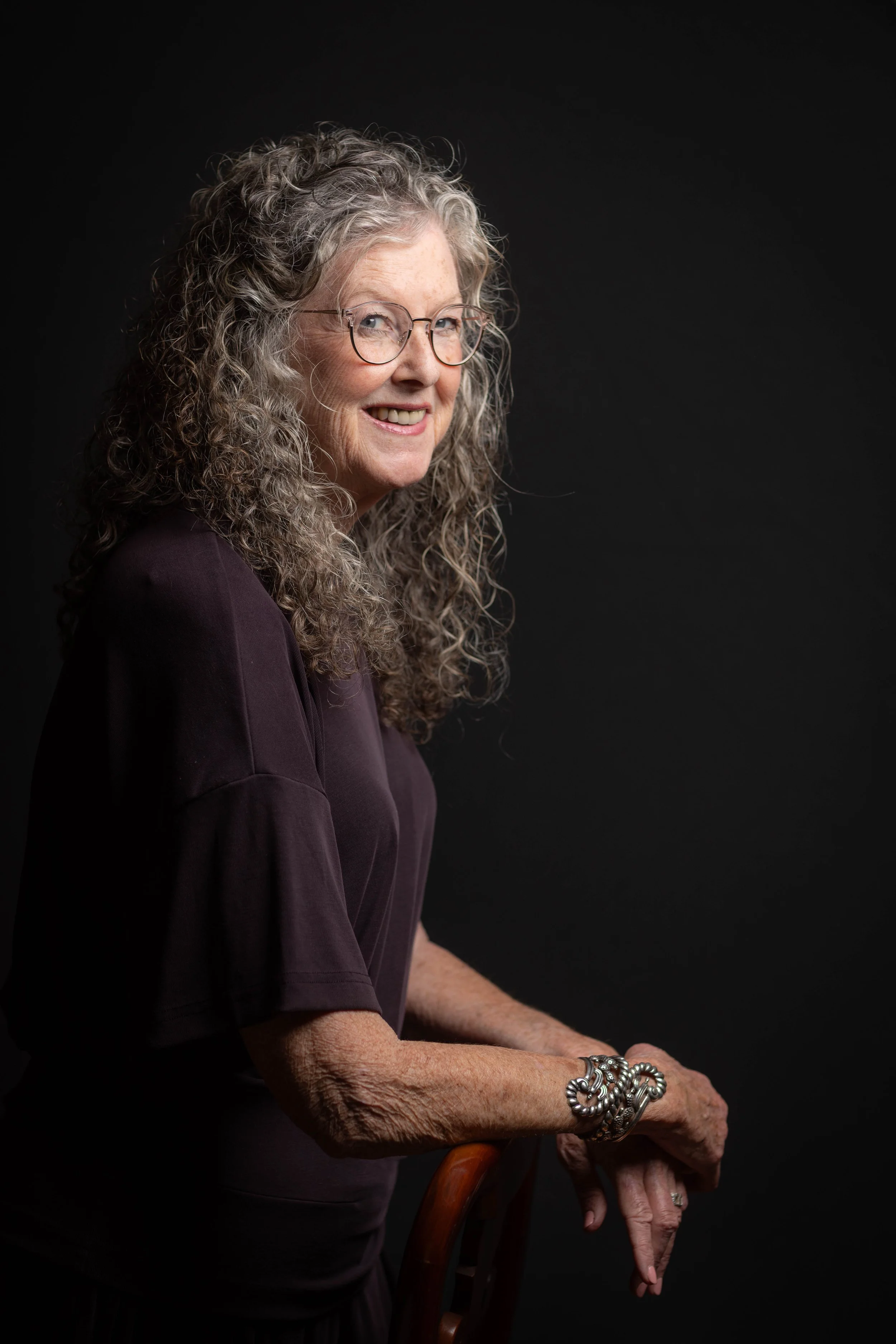 Portrait of author, Kathleen Bethel, with gray, curly hair and glasses, smiling, wearing a dark purple top, jewelry, and holding a cane, against a black background.