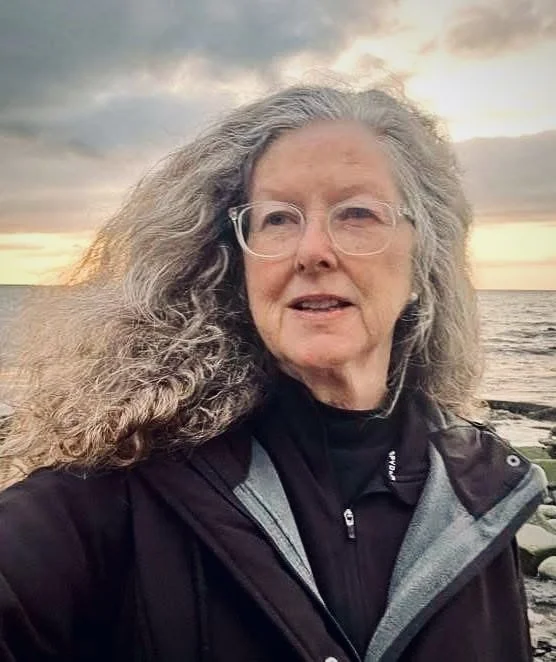 Author, Kathleen Bethel, standing outdoors near the water at sunset, wearing a dark jacket with a gray hood.