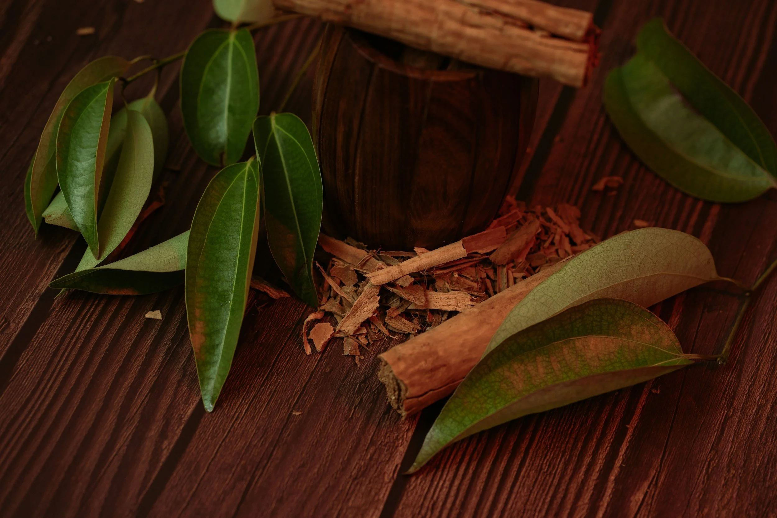 A wooden table with green leaves, a cinnamon stick, and wood shavings surrounding a dark wooden cup or container.