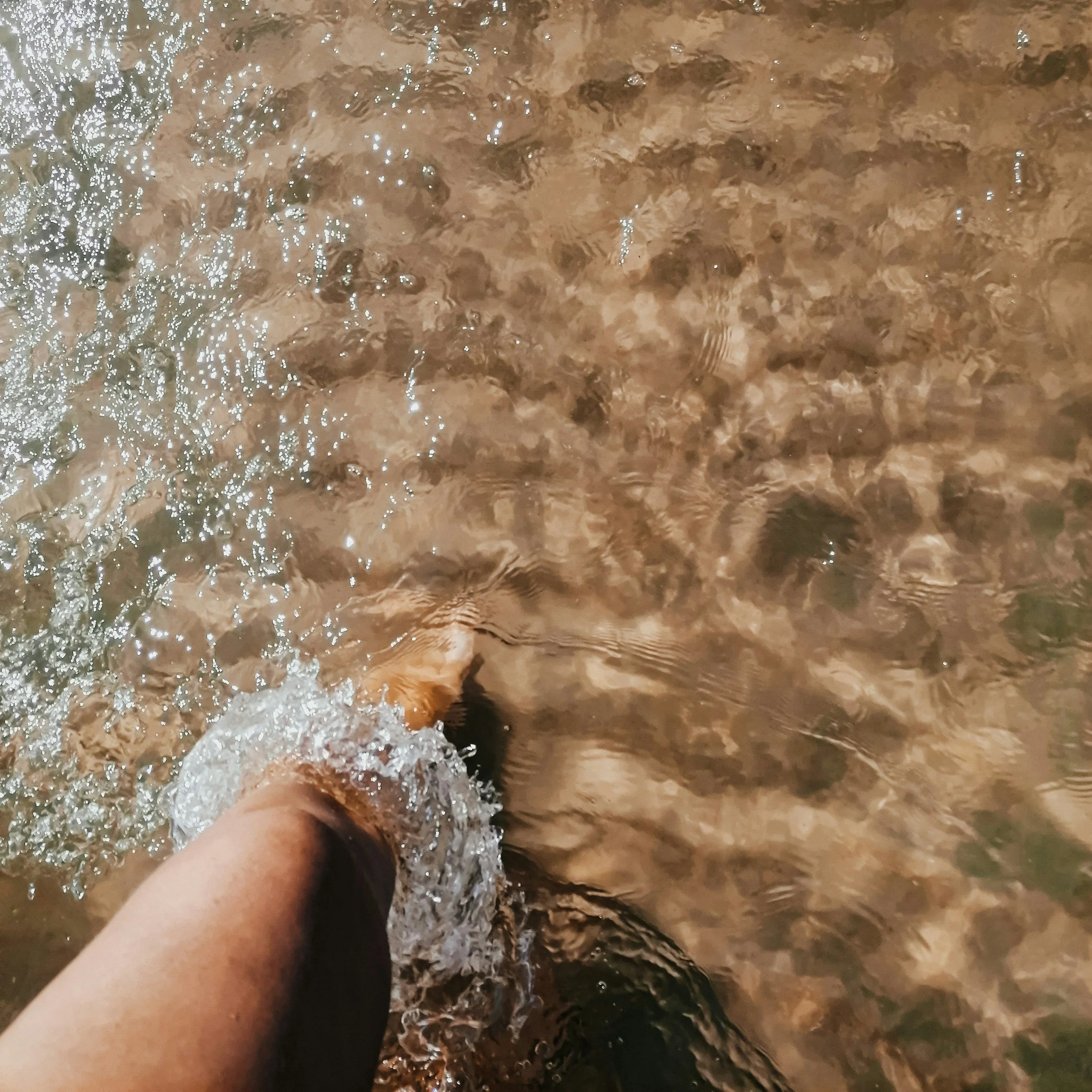 A person's feet walking through shallow, clear water on a sandy beach or riverbed.
