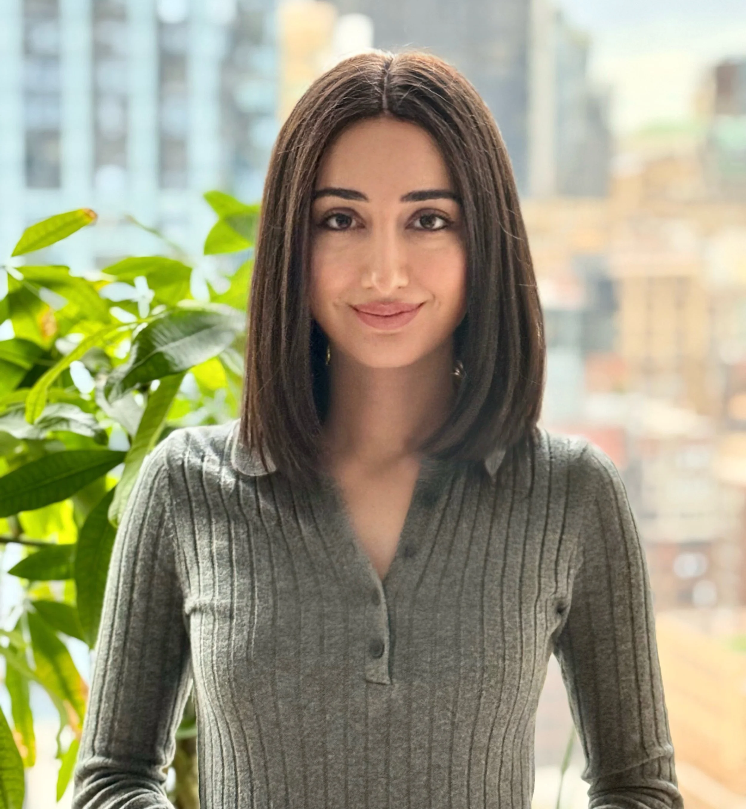 A woman with shoulder-length dark hair, wearing a gray ribbed sweater, standing indoors near a window with green houseplants and a cityscape in the background.