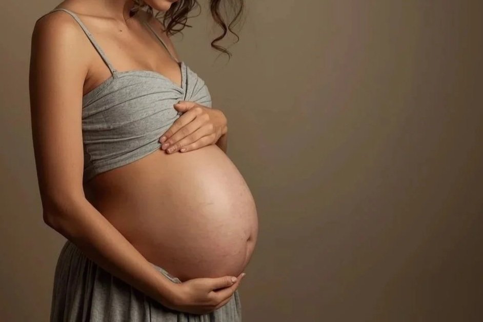 Pregnant woman holding her belly, wearing a gray top and skirt, with a neutral background.