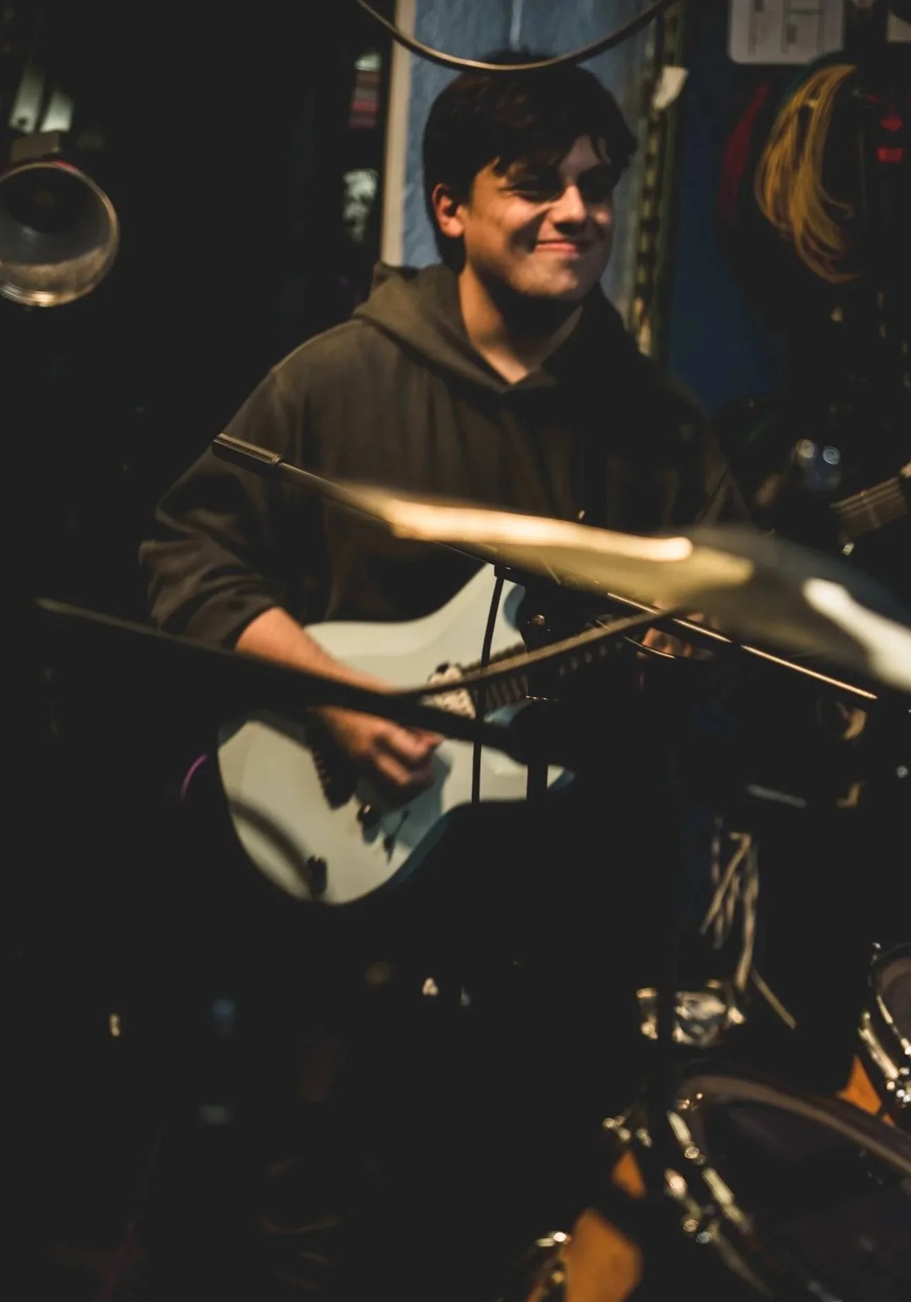 A young man with dark hair smiling while playing an acoustic guitar in a dimly lit room with musical equipment around.