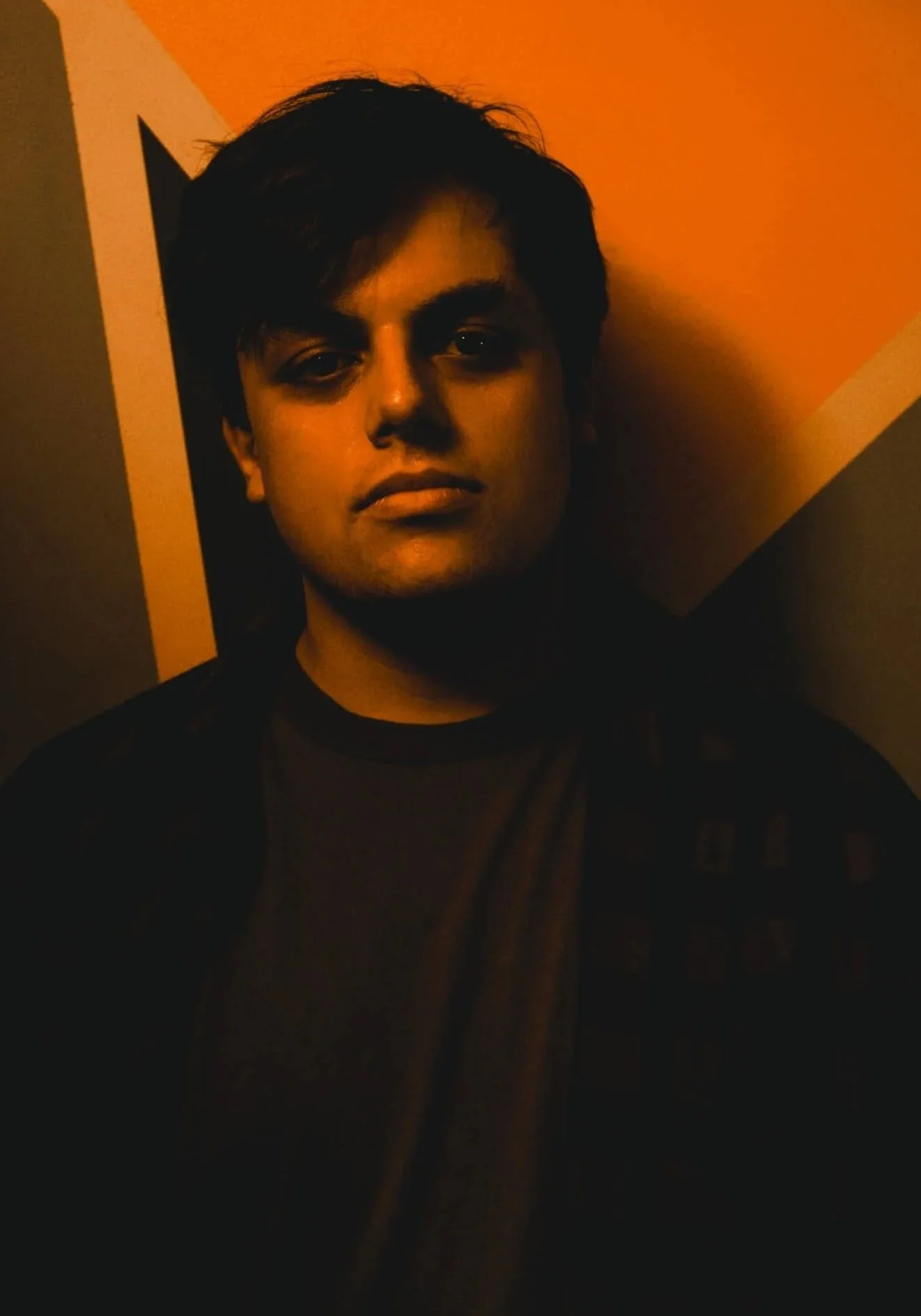 A young man with dark hair posing against a wall with orange, yellow, and gray geometric shapes.