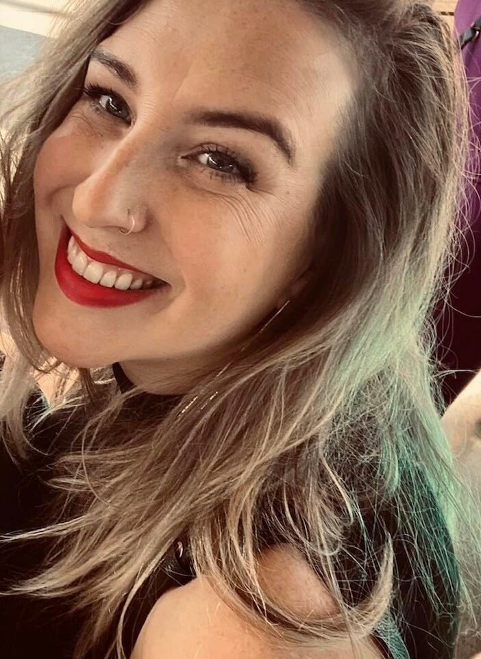 Close-up of a smiling woman with light brown hair, red lipstick, and a nose piercing.