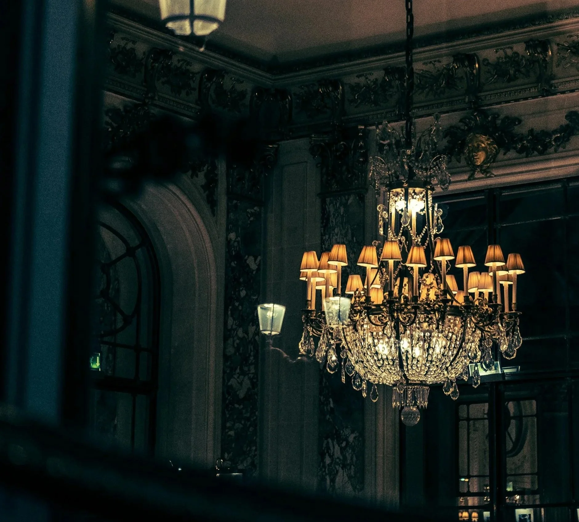 Elegant chandelier with small lampshades hanging in a dimly lit room with ornate moldings and wall sconces.