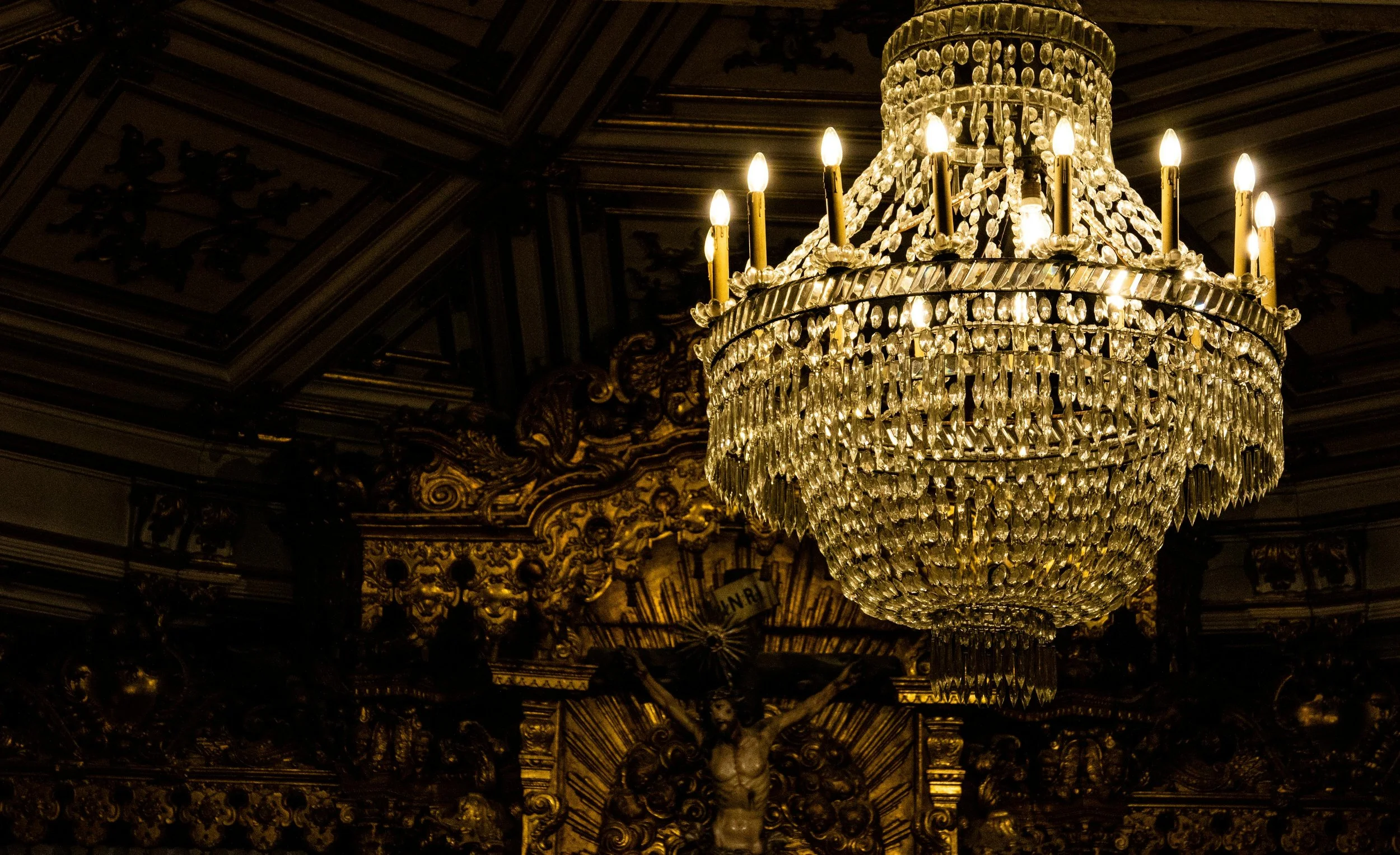 A grand crystal chandelier hanging from an ornate, gilded ceiling in a luxurious, historic interior.