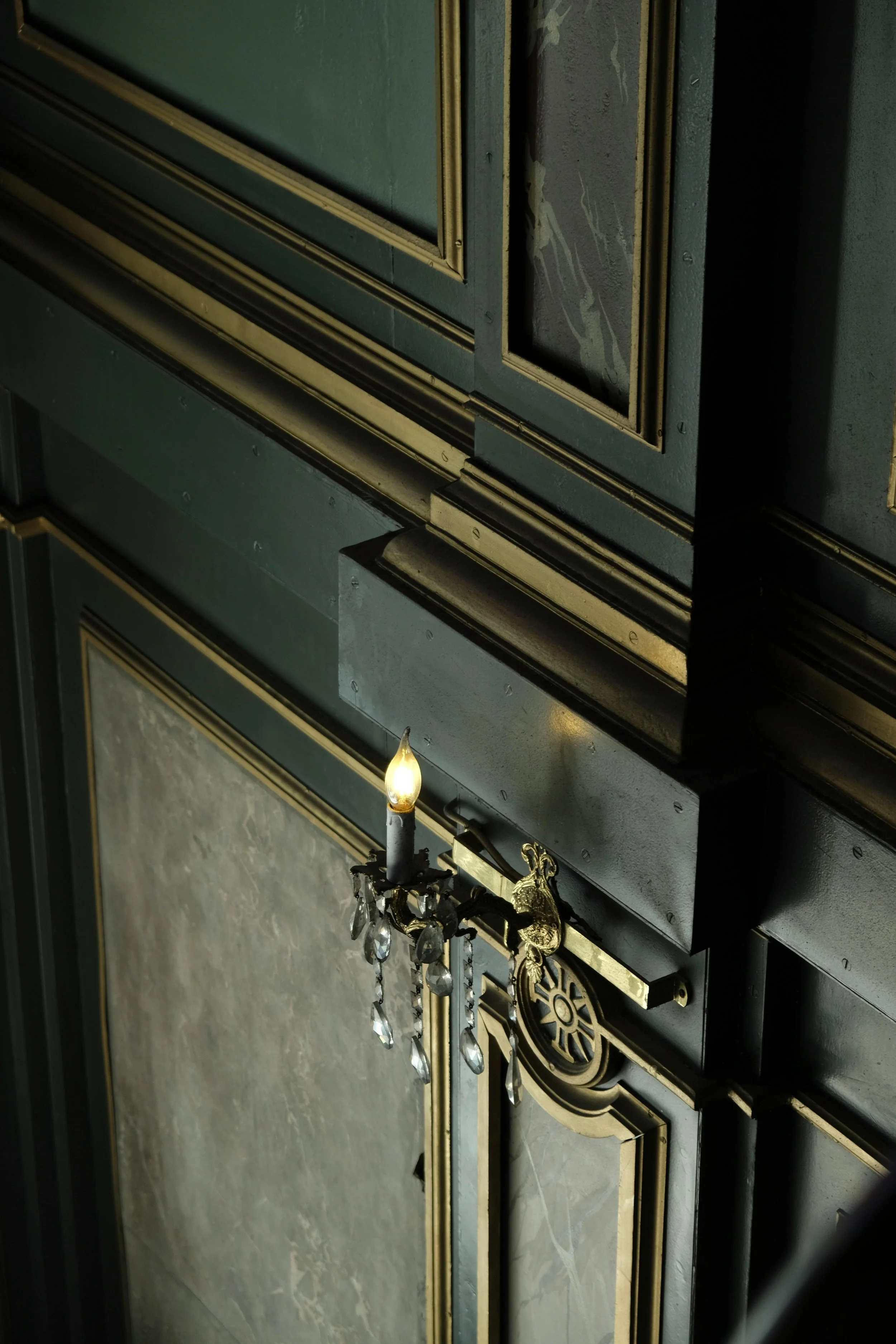 Close-up of a decorative, dark wood cabinet with gold trim. A small chandelier with a candle-shaped light bulb and hanging crystals is attached to the cabinet door.