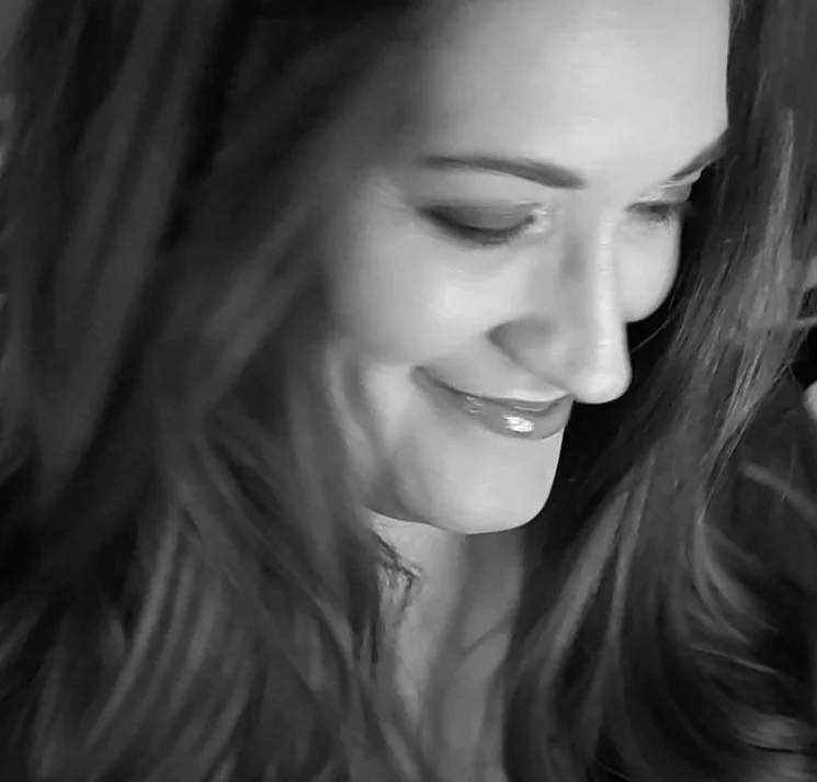 Close-up black and white photo of a woman with long wavy hair smiling with her eyes closed.