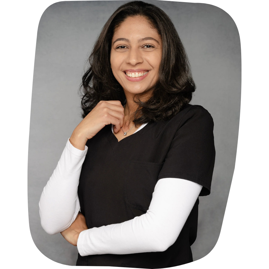 Young woman with dark brown hair smiling, wearing a black scrub top over a white long-sleeve shirt, standing against a gray background.