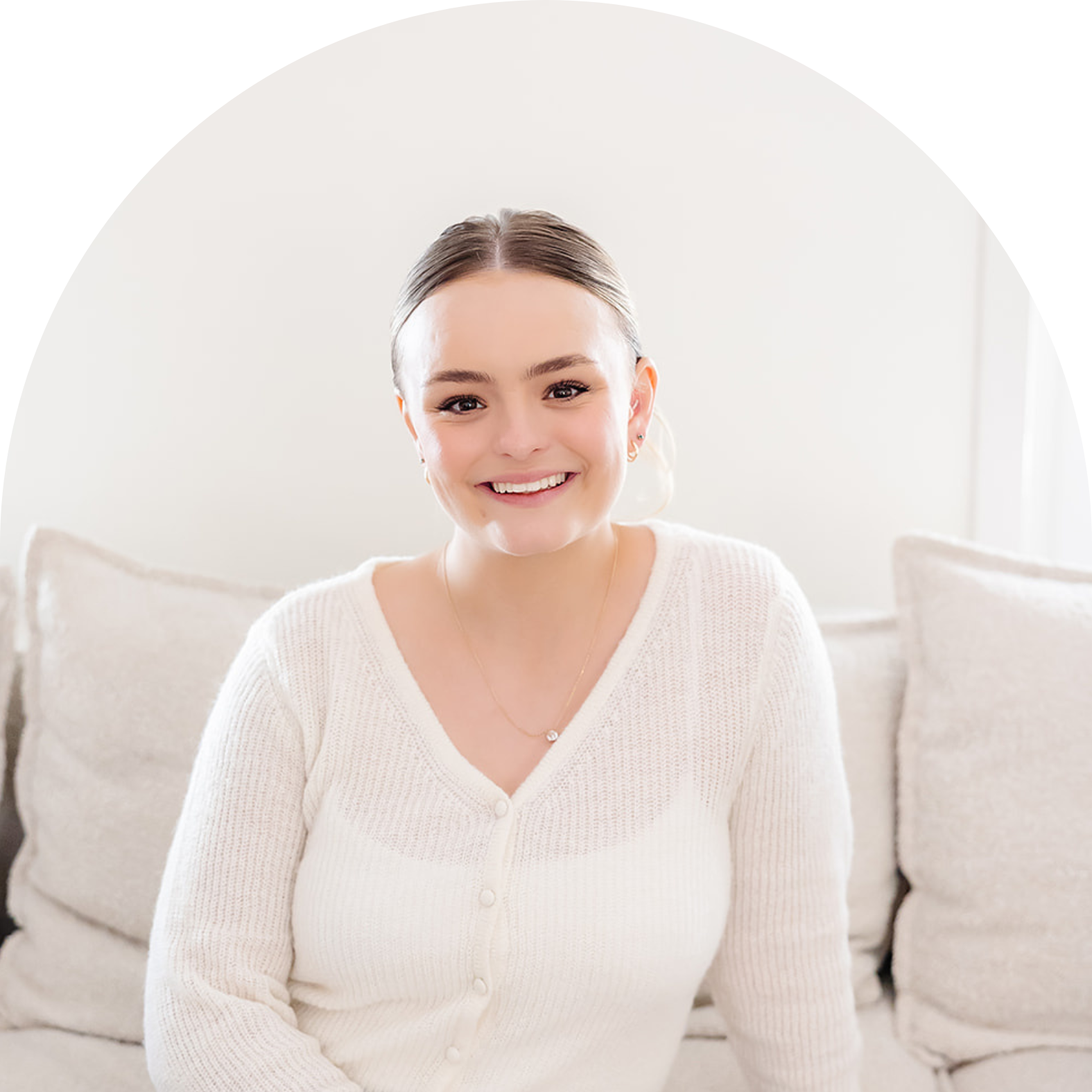 A smiling woman with brown hair, wearing a white cardigan, sitting on a beige couch in a bright, minimalist living room.