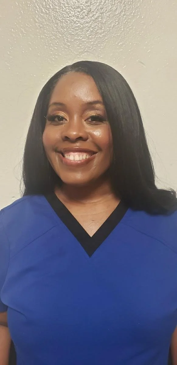 A woman with long black hair and a bright smile is wearing a blue shirt with a black V-neck trim. She is standing against a plain off-white wall.
