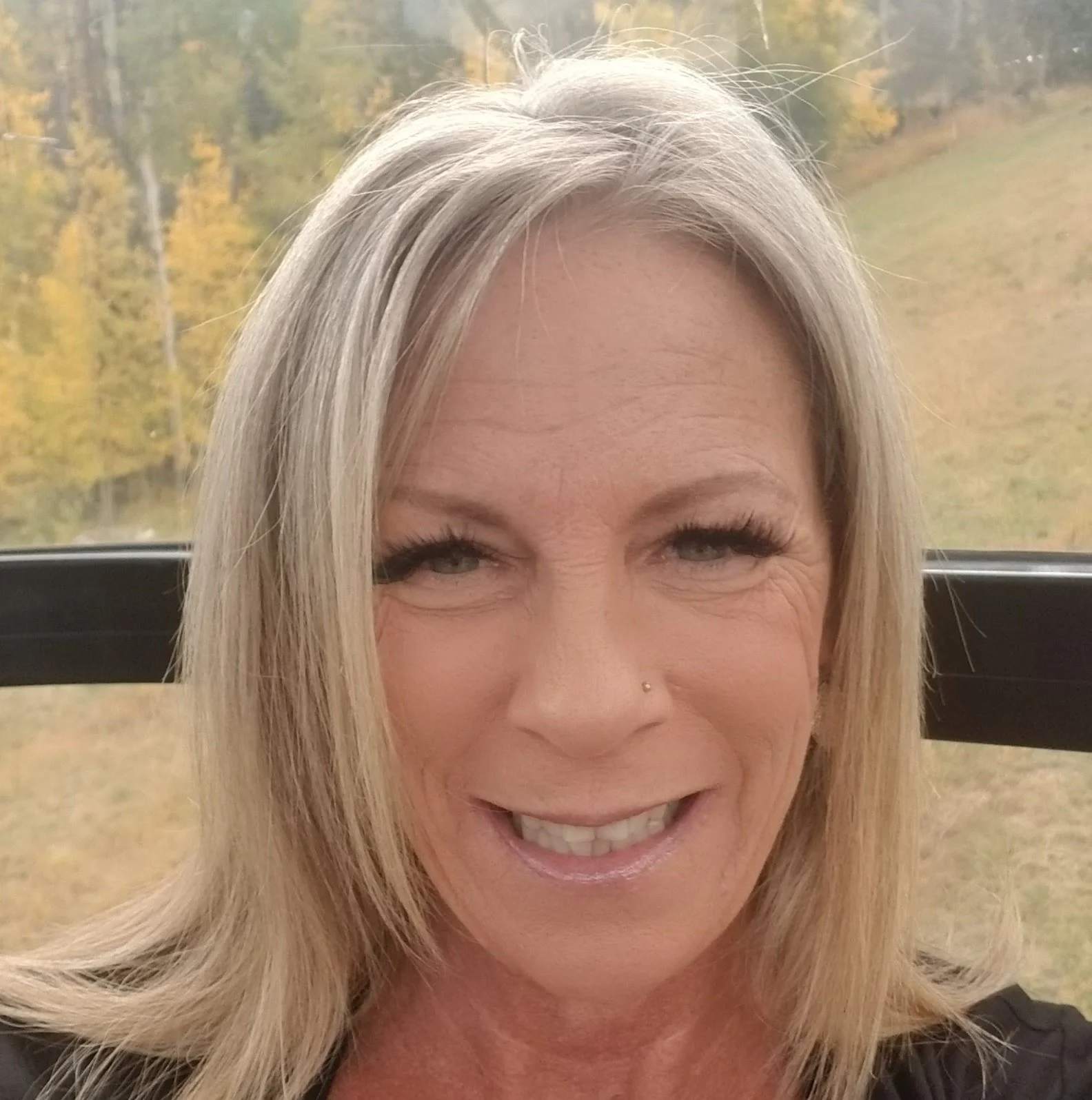 Close-up of a smiling middle-aged woman with blonde hair and a nose piercing, outdoors with a background of yellow and green trees.